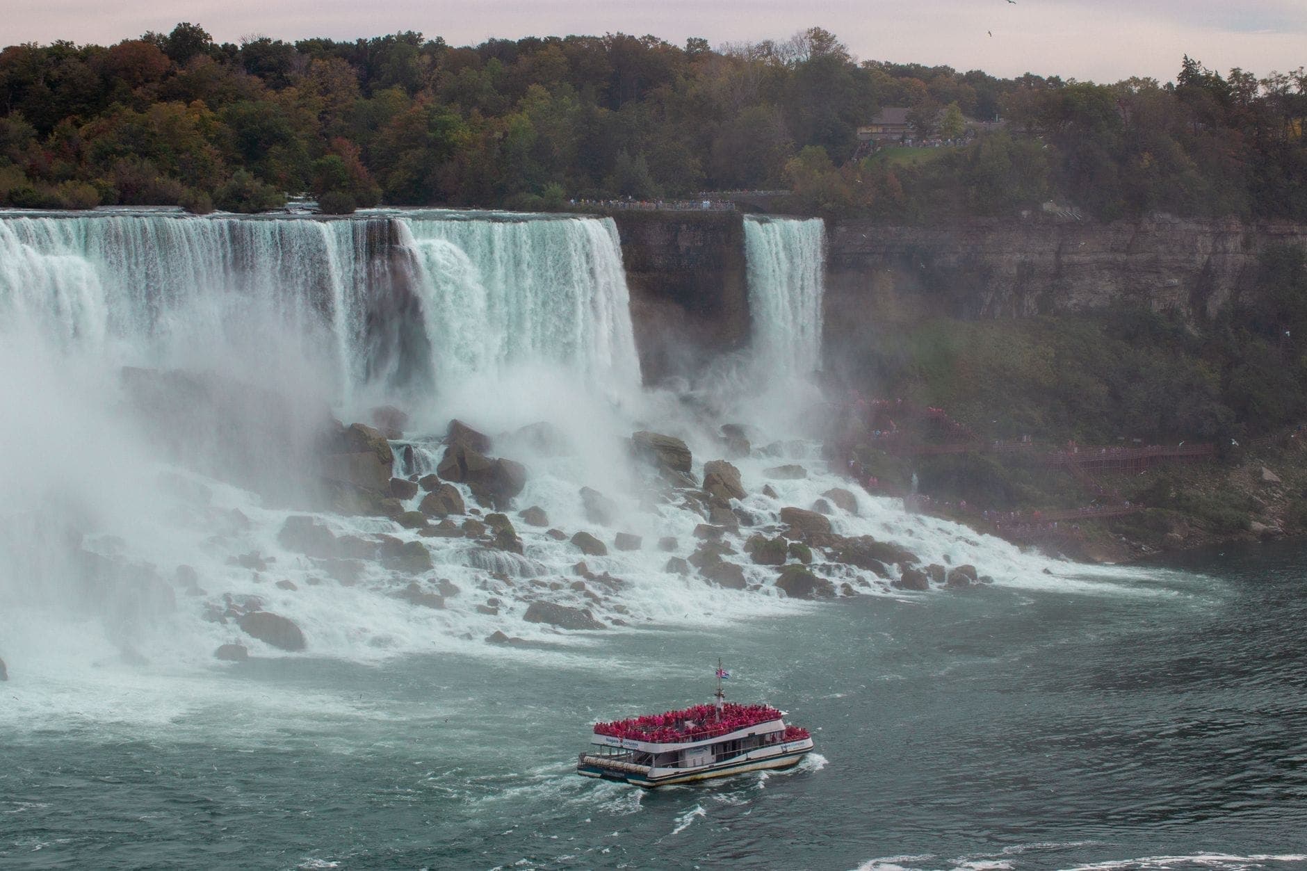 Pemandangan luas Niagara Falls dengan kapal wisata penuh turis mendekati dasar air terjun dan kabut tebal mengepul, dikelilingi pepohonan hijau.