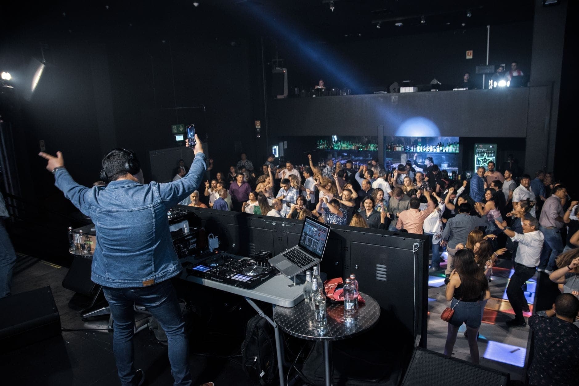 DJ tampil di depan kerumunan penuh yang menari dan bersorak, dikelilingi cahaya berwarna-warni dalam suasana malam yang meriah.