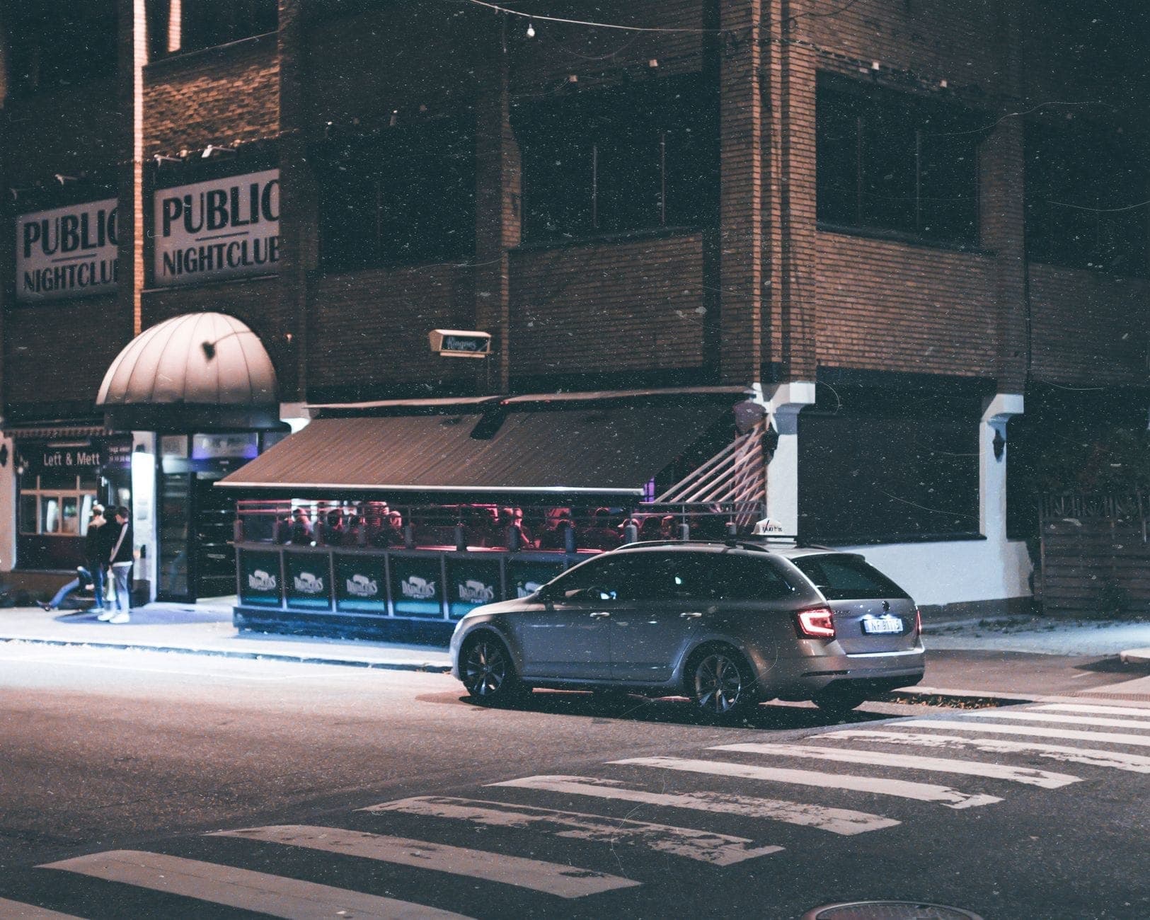 Vista nocturna del exterior del Public Nightclub en Toronto con un auto oscuro estacionado al frente y las luces de la ciudad iluminando la escena.