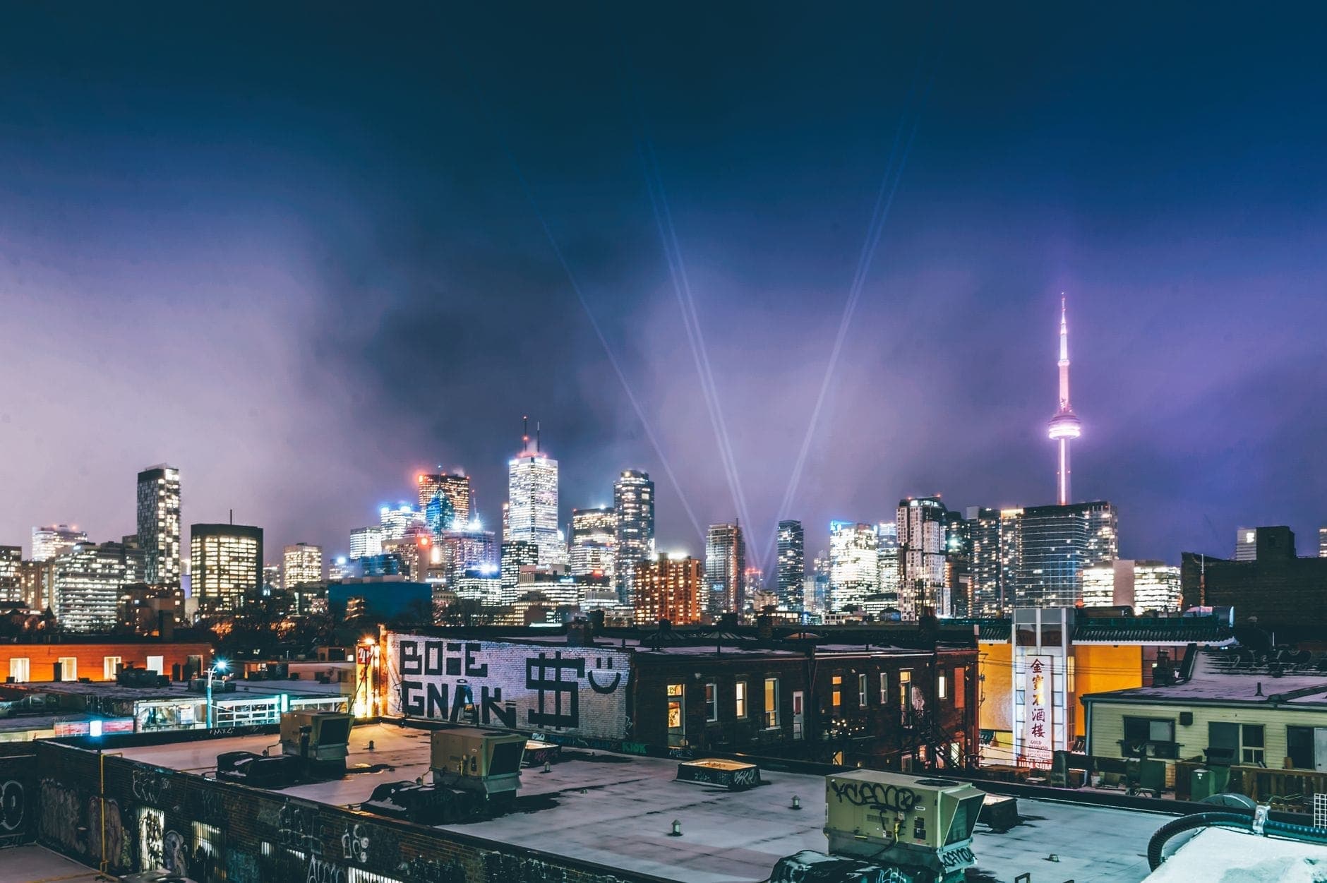 Horizonte de Toronto de noche con brillantes luces de la ciudad, la CN Tower iluminada y haces de luz en el cielo sobre los tejados.