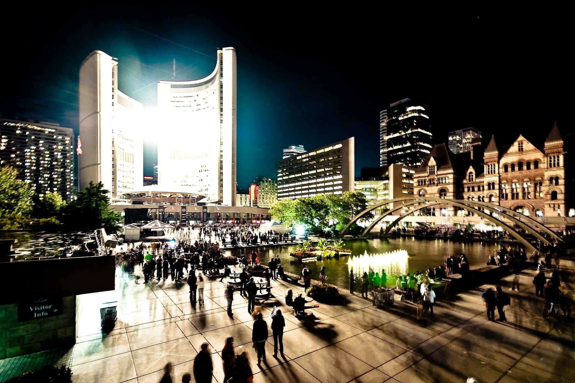 Kerumunan berkumpul di Nathan Phillips Square Toronto pada malam hari, dengan City Hall yang terang benderang dan kolam pantulan yang dikelilingi bangunan modern dan bersejarah.