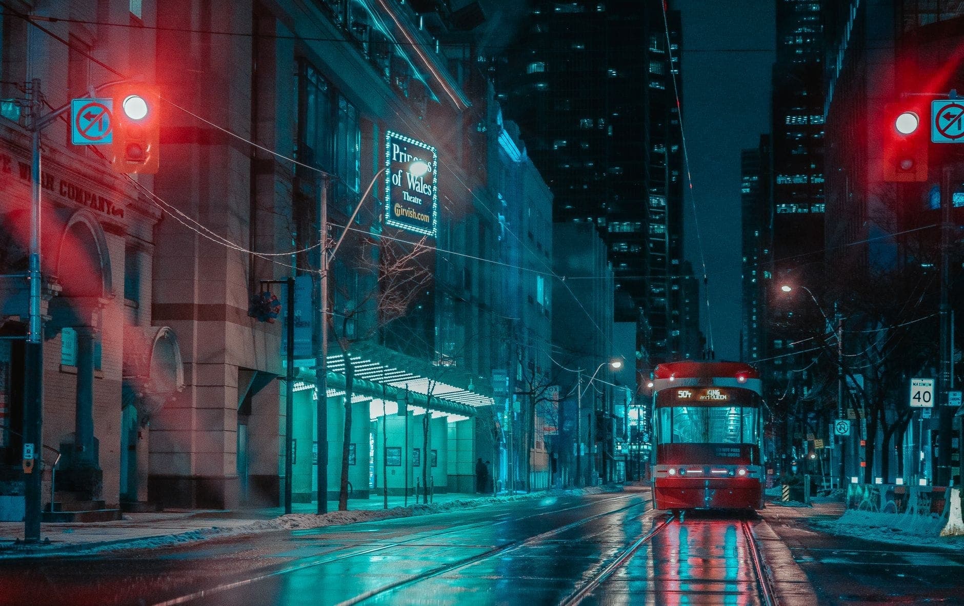 Un tram di Toronto percorre una strada cittadina bagnata e illuminata al neon di notte, circondata da alti edifici e semafori luminosi.