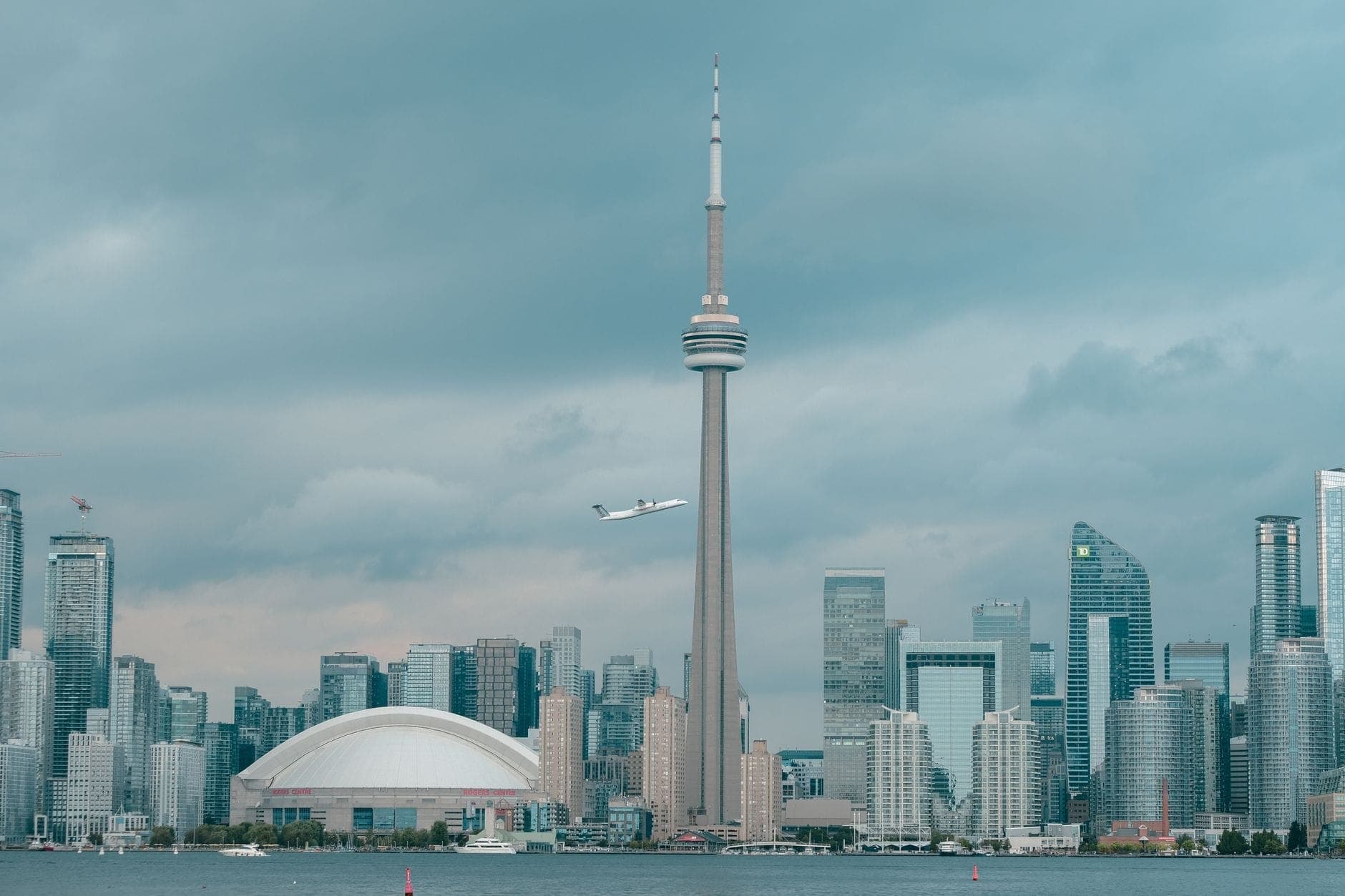Cakrawala Toronto dengan CN Tower dan Rogers Centre di bawah langit dramatis, dilihat dari seberang perairan dengan pesawat terbang di atasnya.