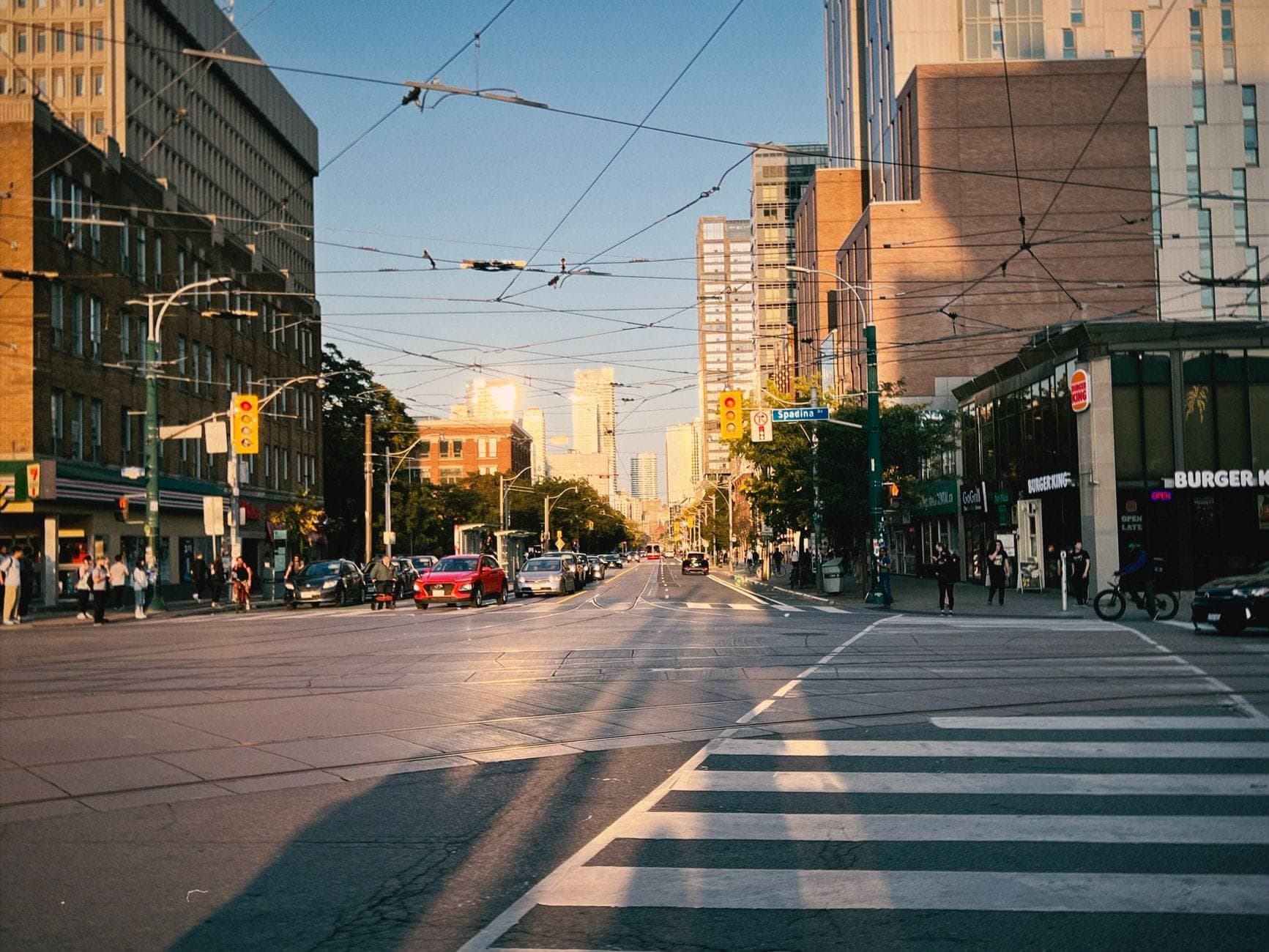 Ampia veduta di una trafficata strada di Toronto con ristoranti e locali da asporto lungo il marciapiede, cavi della luce in alto e persone che camminano nel tardo pomeriggio.