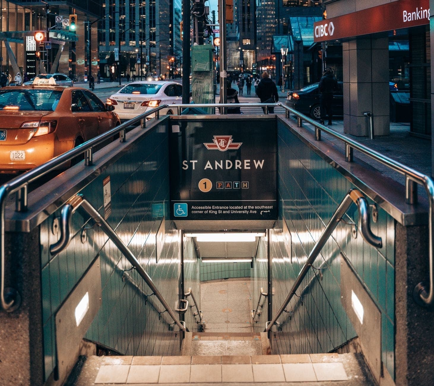 Pintu masuk stasiun subway St. Andrew Toronto dengan tangga menuju bawah tanah, dikelilingi gedung-gedung pusat kota, lalu lintas, dan pejalan kaki di bawah cahaya sore.