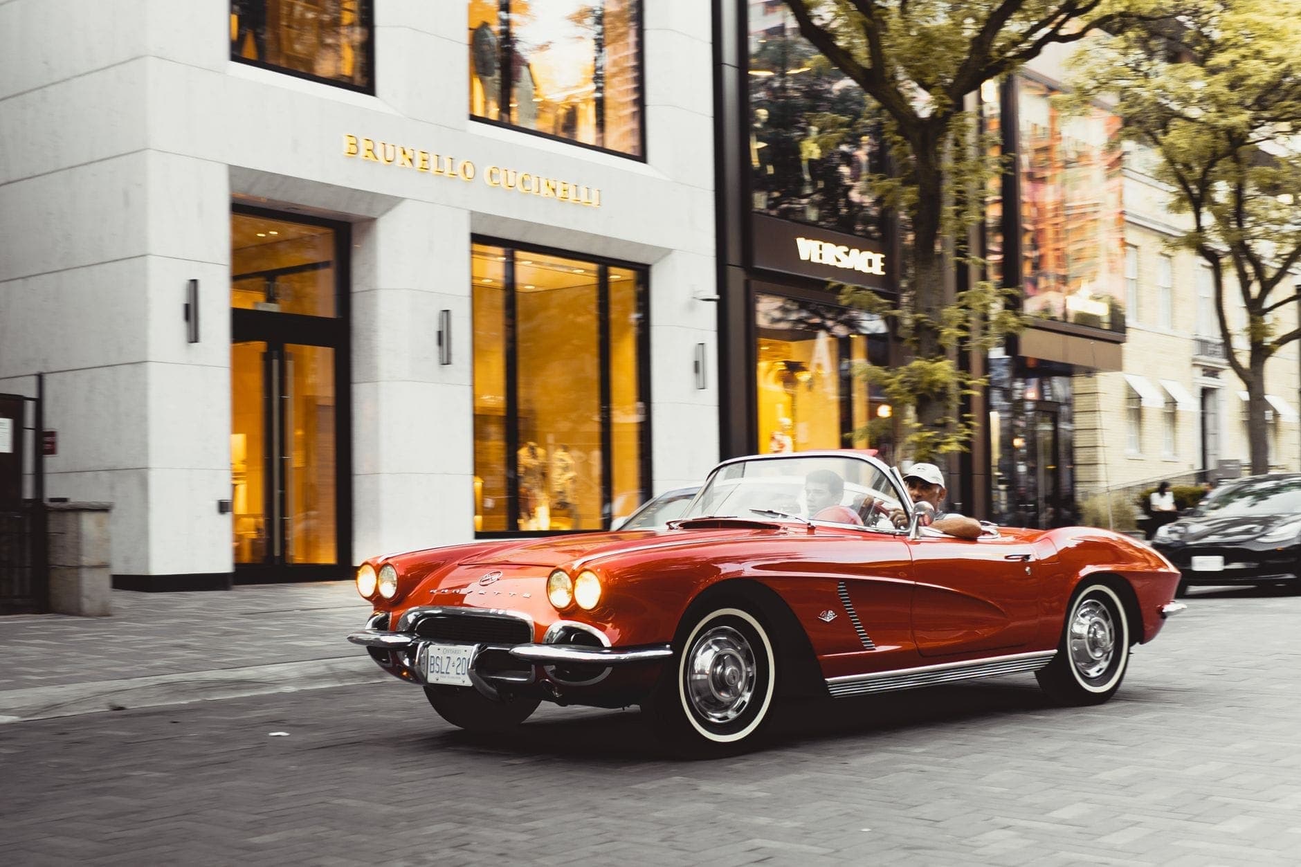Elegantes escaparates de Brunello Cucinelli y Versace en una sofisticada calle comercial con un clásico descapotable rojo pasando por delante.