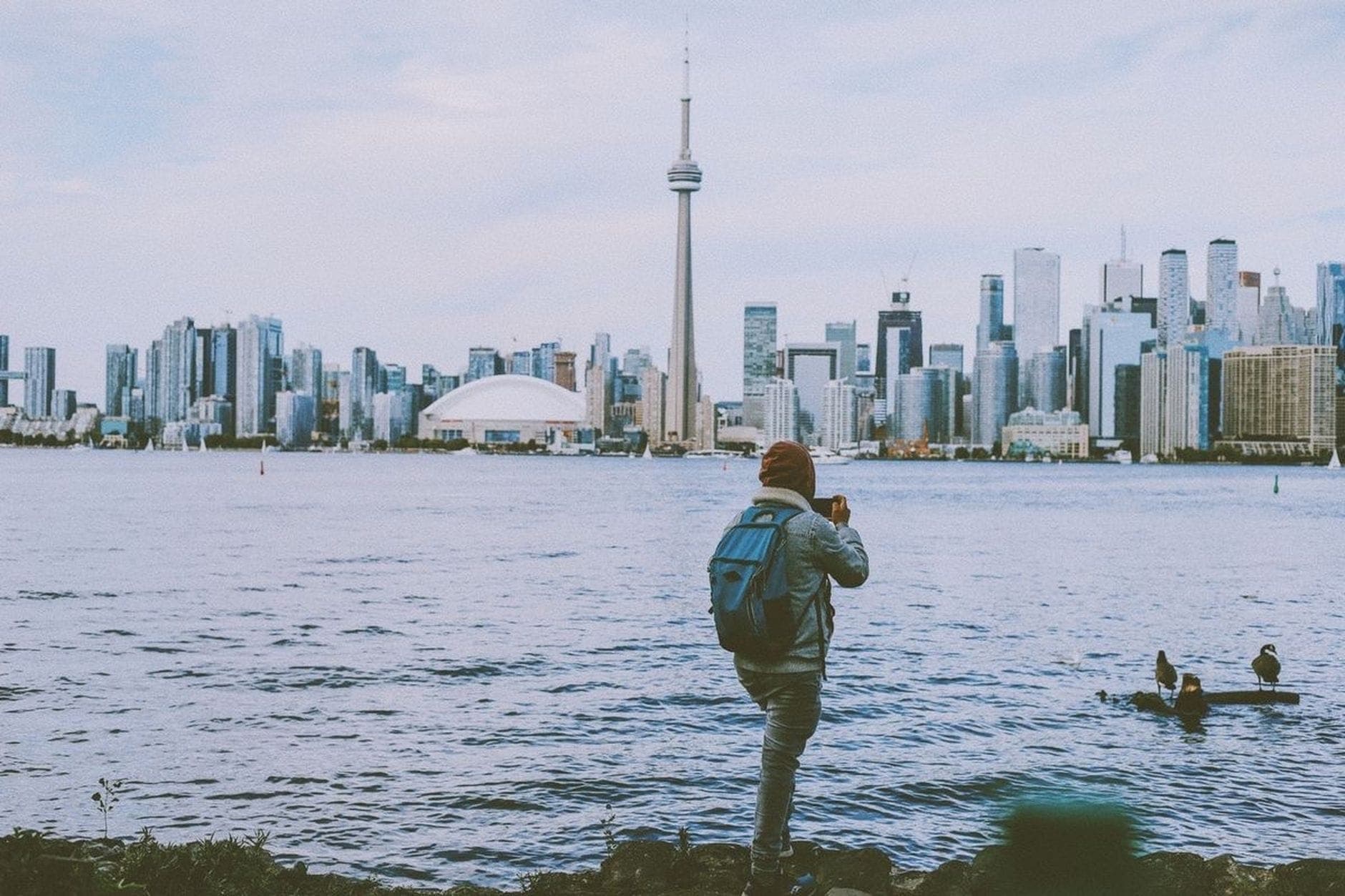 Wisatawan solo dengan ransel memotret cakrawala Toronto dari seberang air, menampilkan CN Tower dan gedung-gedung pusat kota di hari berawan.