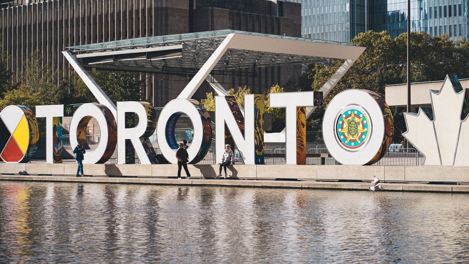 Personas caminando frente al icónico letrero de Toronto en Nathan Phillips Square, con edificios modernos al fondo.