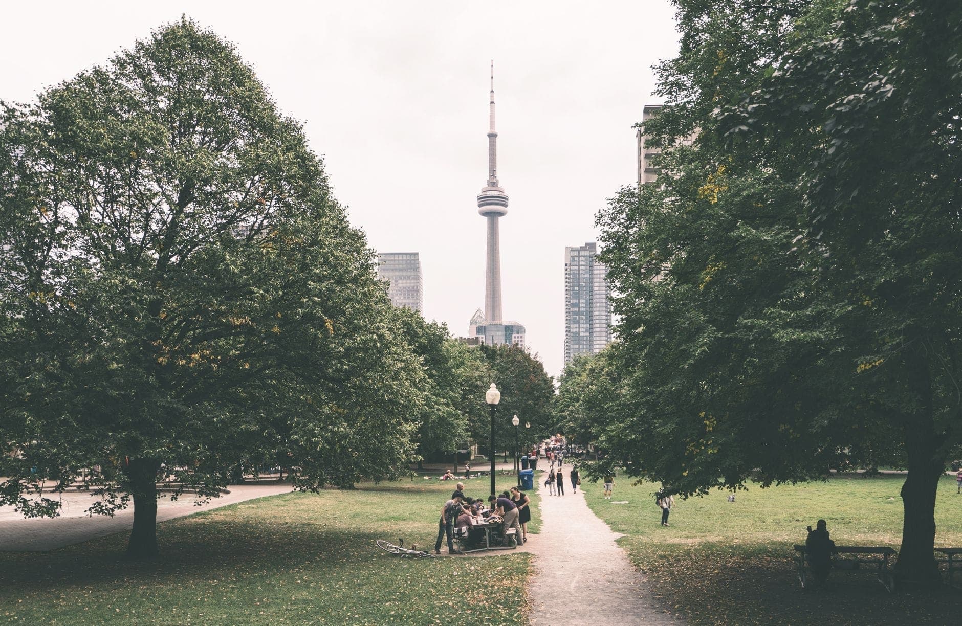 Orang-orang berjalan di jalur taman menuju CN Tower Toronto, dikelilingi pepohonan hijau lebat dan gedung-gedung kota, di hari yang mendung.