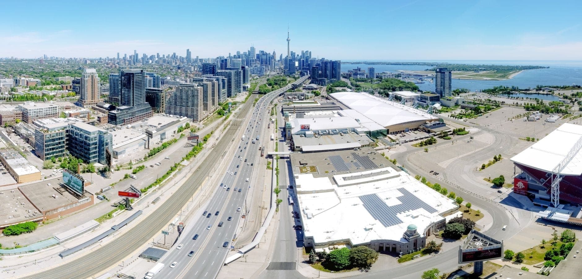 A panoramic aerial view of Toronto's waterfront showing skyline, CN Tower, roadways, lakeshore parks, and waterfront features on a clear day.