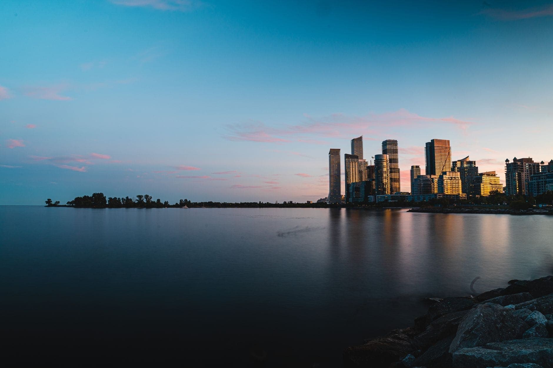 Toronto waterfront view at dusk with high-rise condos, parkland, and calm lake reflecting the sky, illustrating east-west waterfront contrast.
