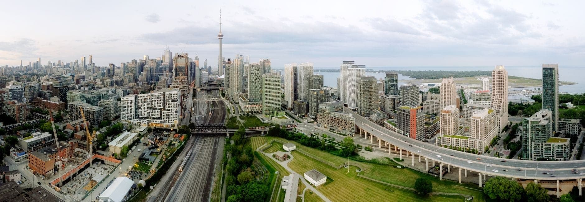 Wide aerial view of Toronto waterfront, showing expressway, green park spaces, lakeshore, and skyline including the CN Tower and high-rise buildings.