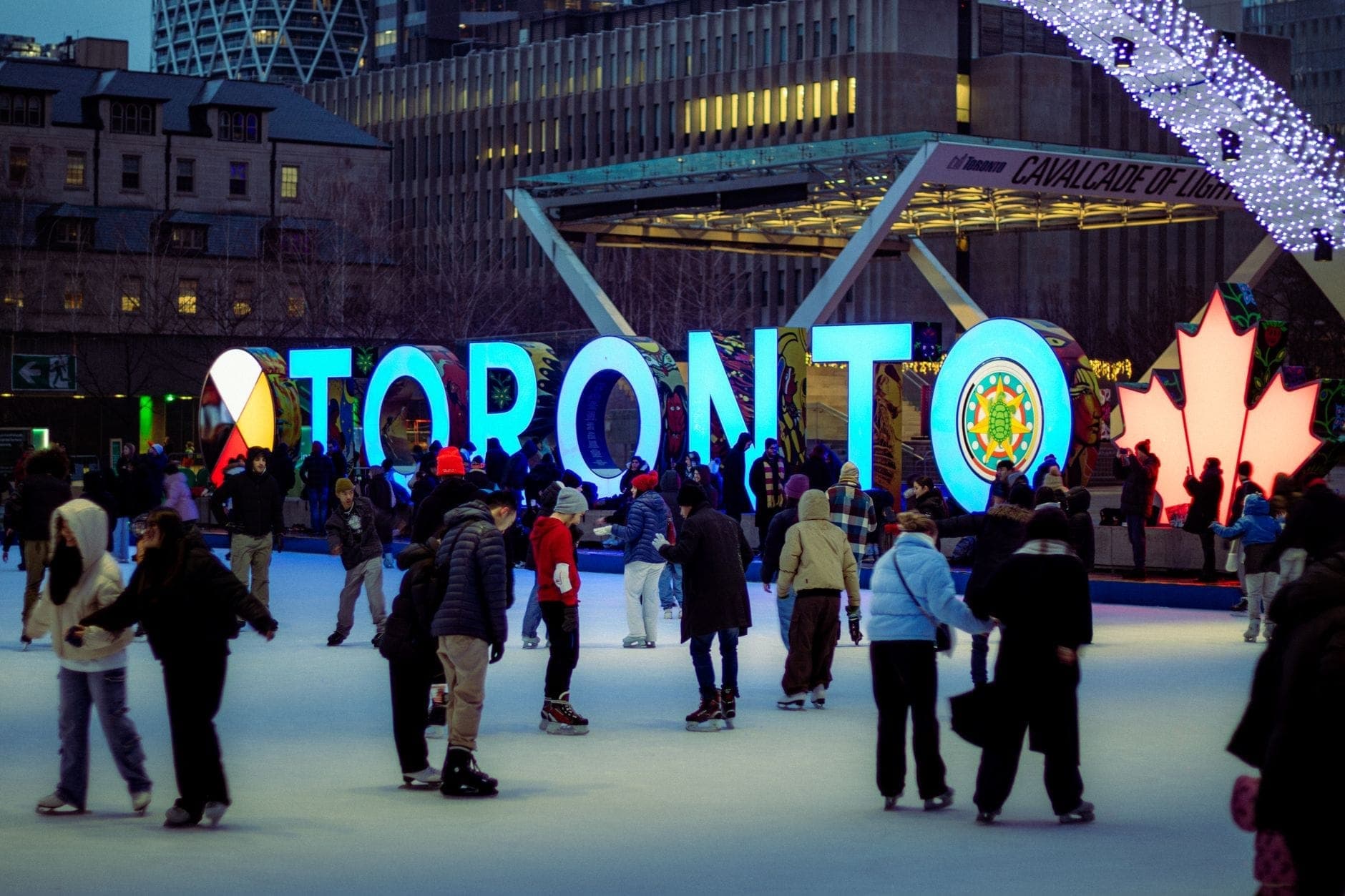 Orang-orang dari berbagai usia bermain seluncur es di Nathan Phillips Square, di depan papan nama Toronto yang bercahaya, dengan gedung-gedung kota di latar belakang.