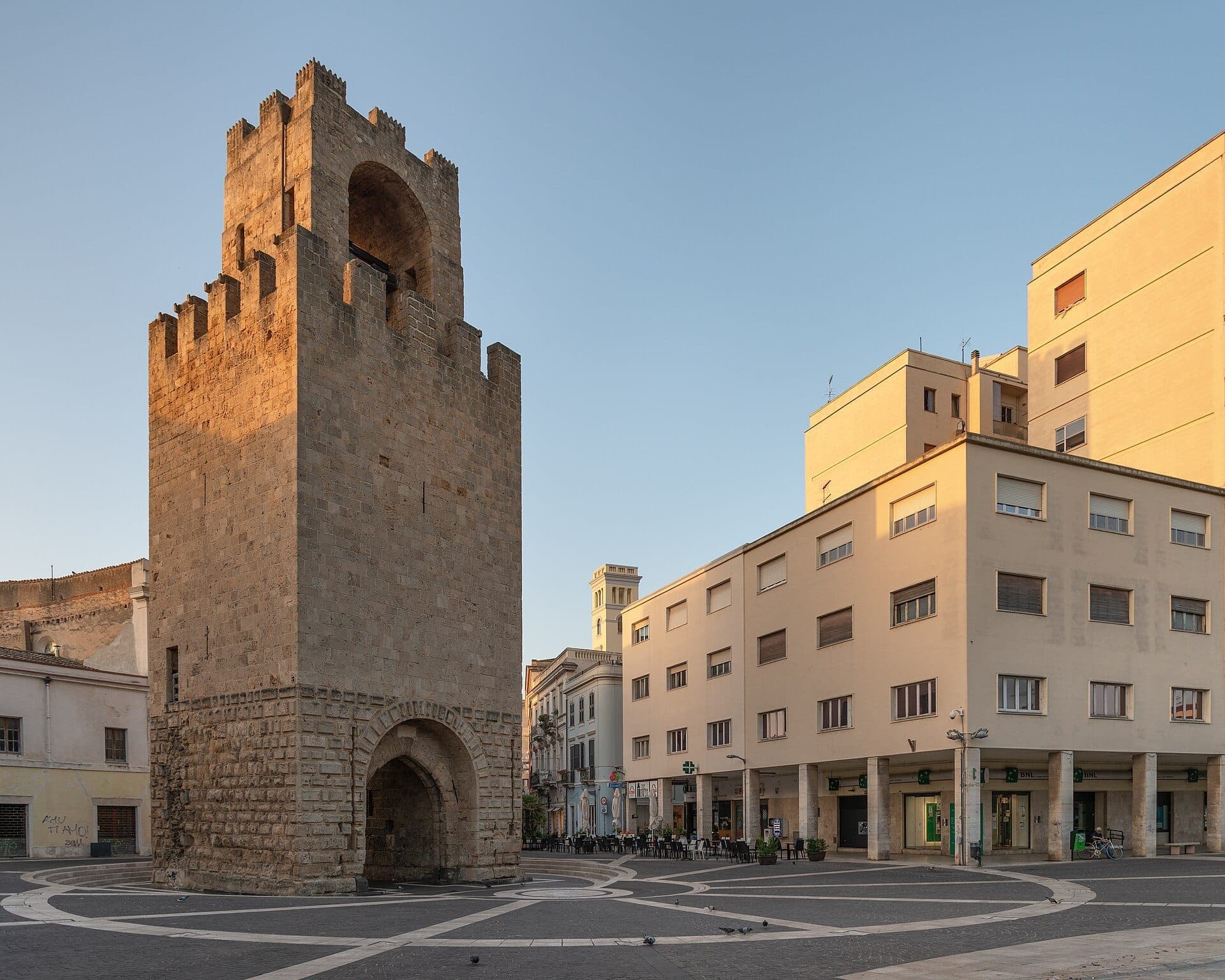 La Torre di Mariano II se alza imponente en la Piazza Roma de Oristano, rodeada de edificios modernos bajo la clara luz del atardecer.