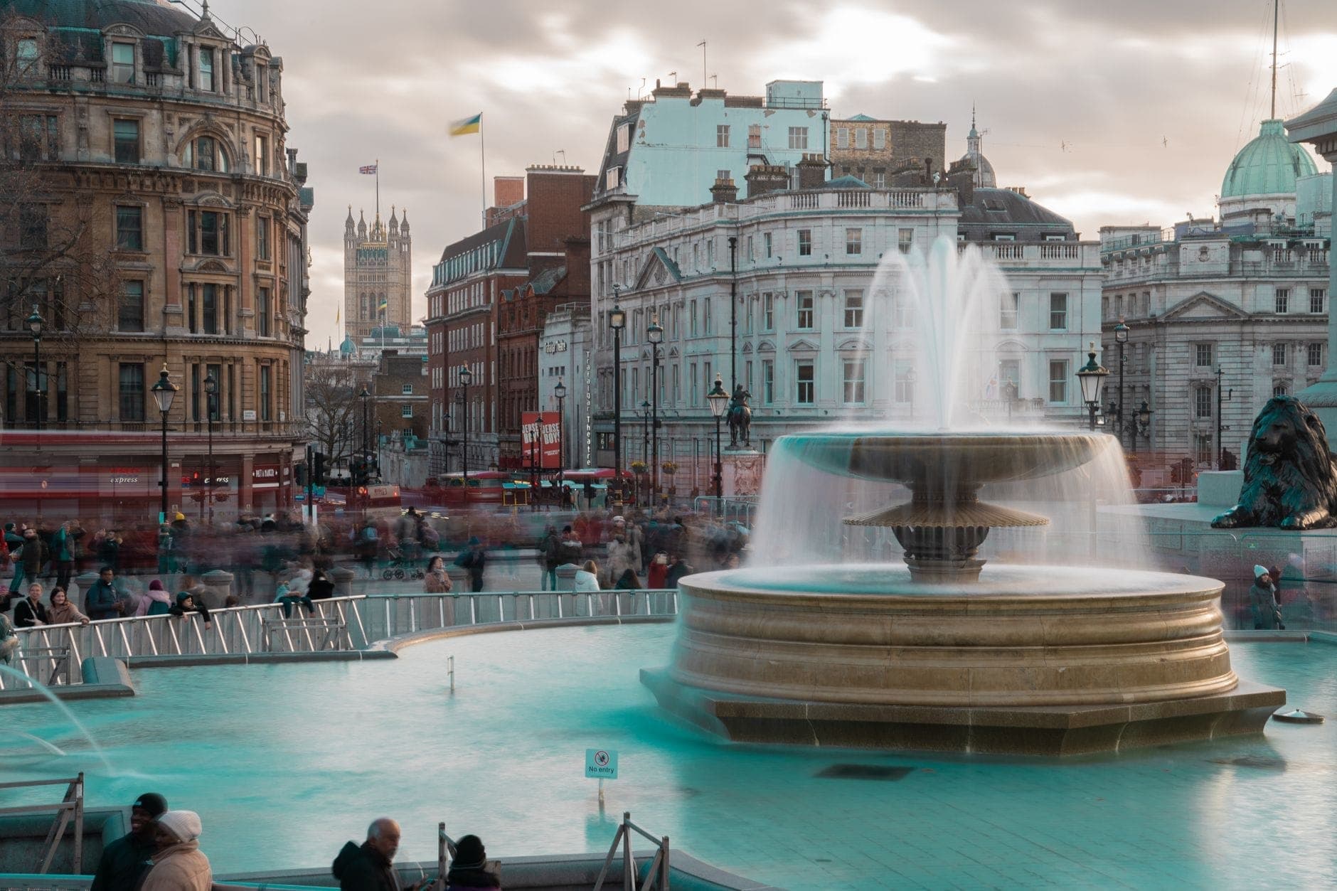 View of Trafalgar Square showing the central fountain, surrounding historic buildings, and crowds of people on a cloudy day in London.