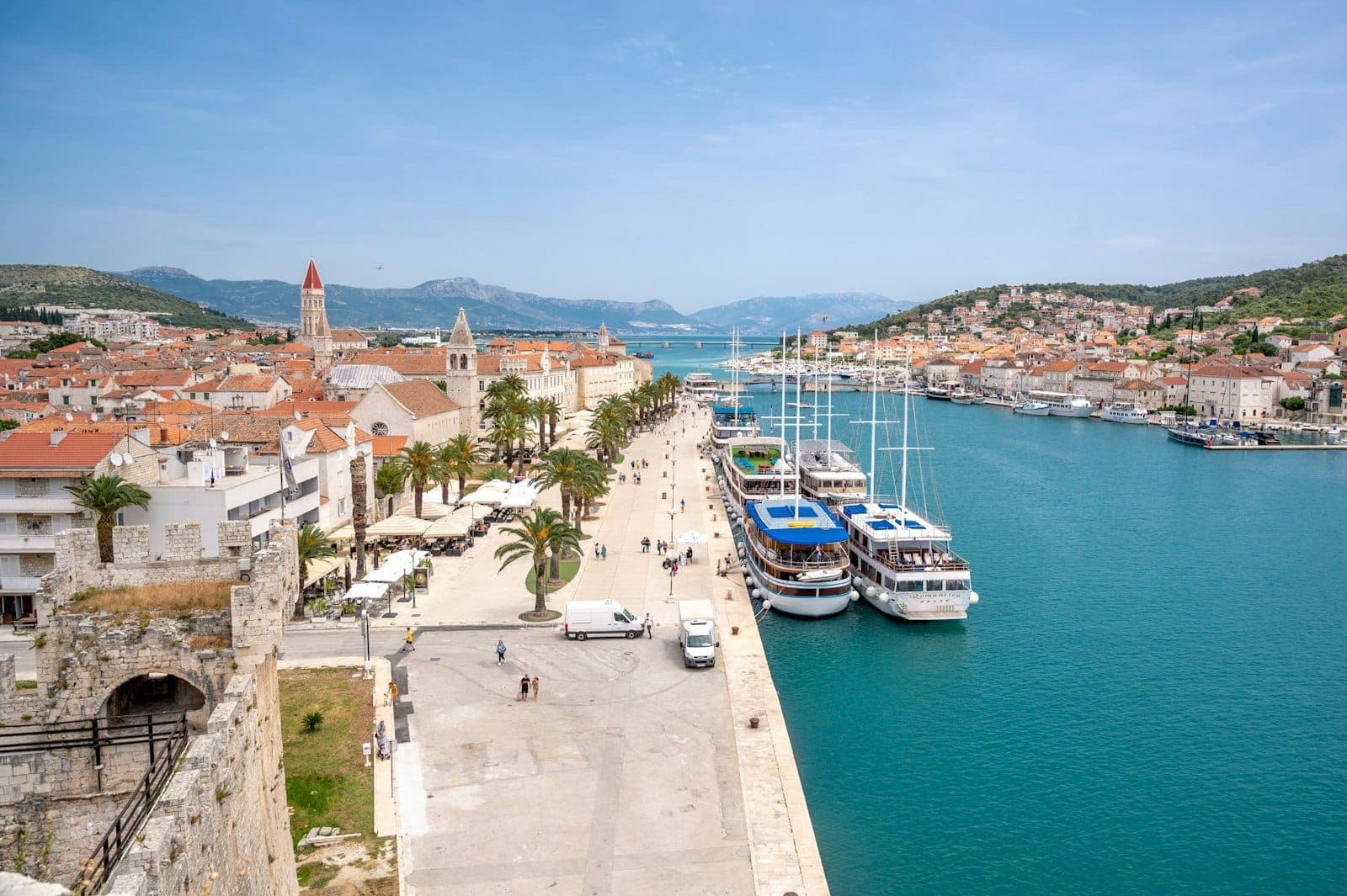 Vista sul lungomare storico di Trogir con edifici in pietra, passeggiata fiancheggiata da palme, barche ormeggiate e acqua blu cristallina sotto un cielo soleggiato.