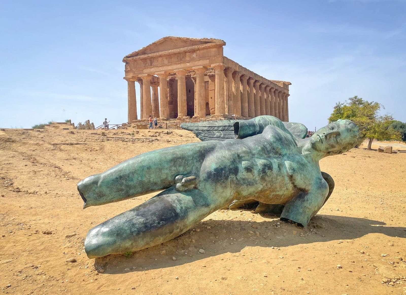 Kuil Yunani kuno di Agrigento dengan patung perunggu besar yang tumbang di latar depan di bawah langit cerah.