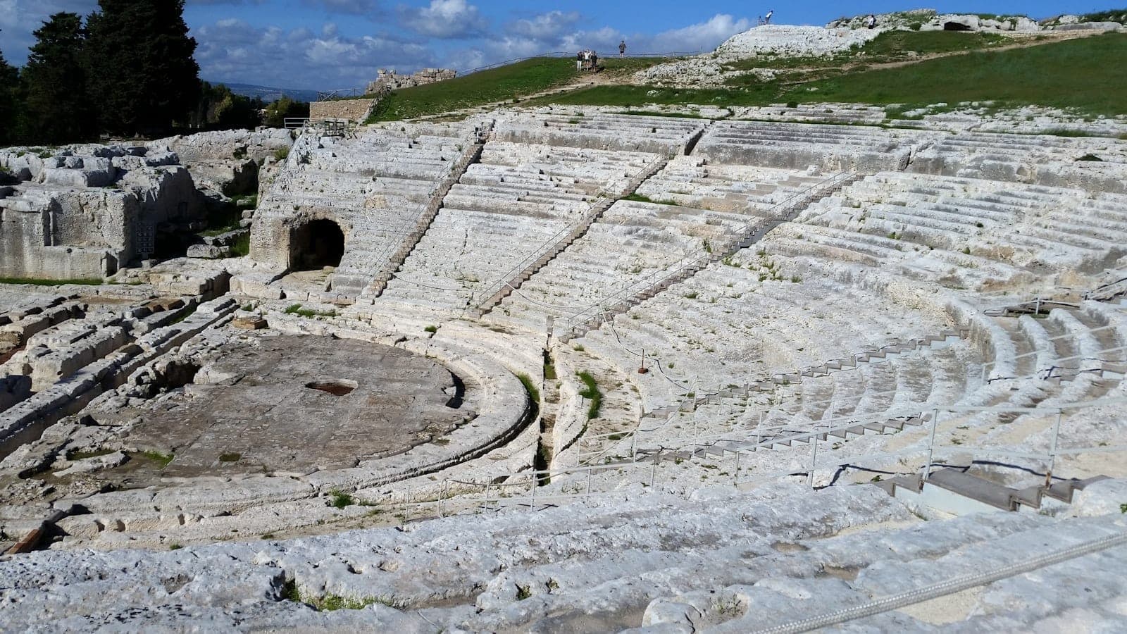Pemandangan luas teater Yunani kuno di Syracuse, Sisilia, dengan tempat duduk batu dan rumput hijau di atasnya.