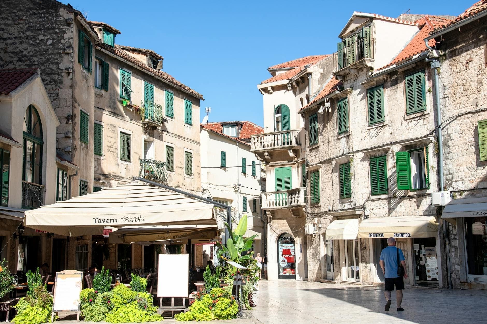Affascinante piazza a Spalato con edifici storici in pietra, persiane verdi, caffè e un pedone, che evoca la vivace scena dell'ospitalità della città.