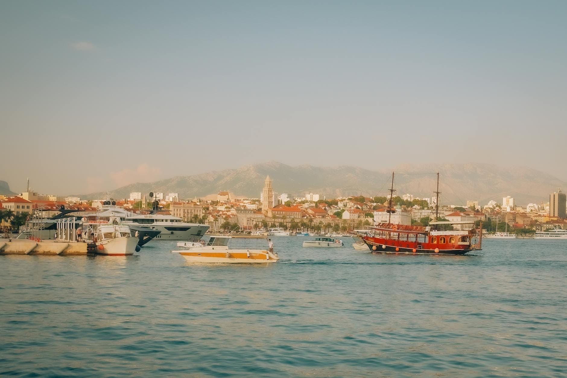 Diverse barche e yacht nel porto di Spalato con il riconoscibile skyline della città vecchia e le montagne in lontananza sotto cieli sereni.