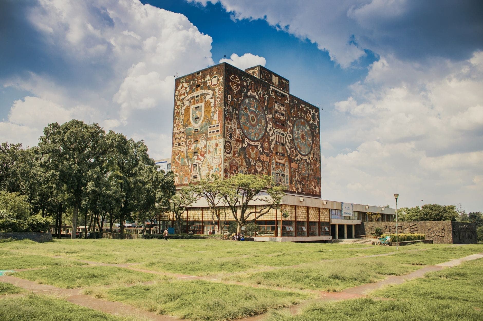 มุมมองกว้างของหอสมุดกลาง UNAM กับภาพจิตรกรรมฝาผนังเด่น พื้นสนามหญ้าและต้นไม้เขียวขจี ใต้ท้องฟ้าที่มีเมฆบางเบาในยามบ่าย