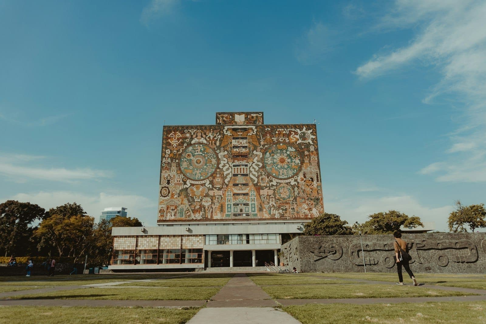 มุมหน้าห้องสมุดกลาง UNAM ที่มีจิตรกรรมฝาผนังชื่อดัง ท้องฟ้าใส สนามหญ้าโล่งและผู้คนเดินผ่านบางตา