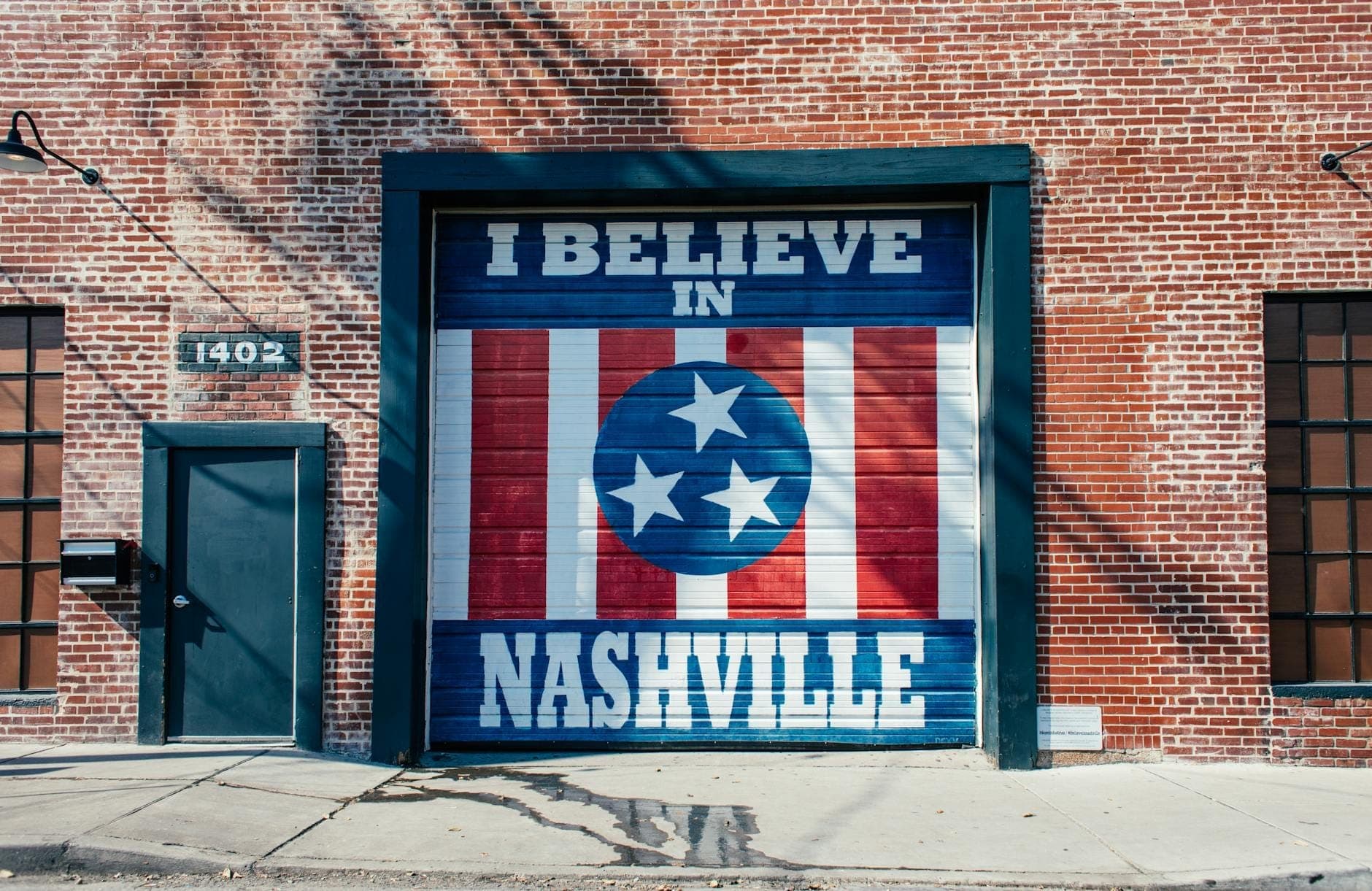 Ein bekanntes 'I Believe in Nashville'-Wandgemälde, das auf eine Garagentür eines roten Backsteingebäudes in einer städtischen Straßenumgebung gemalt wurde.