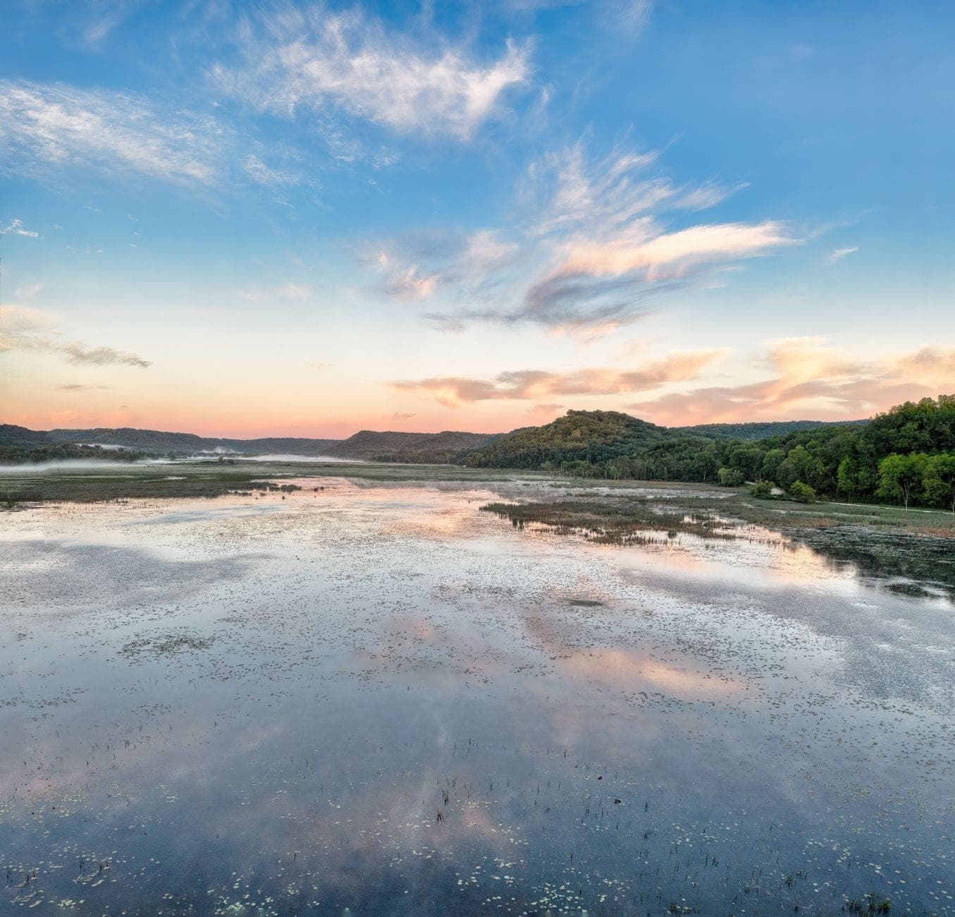 Ein breiter Fluss oder Feuchtgebiet mit spiegelndem Wasser, umgeben von bewaldeten Hügeln bei Sonnenuntergang in der Nähe von Nashville.