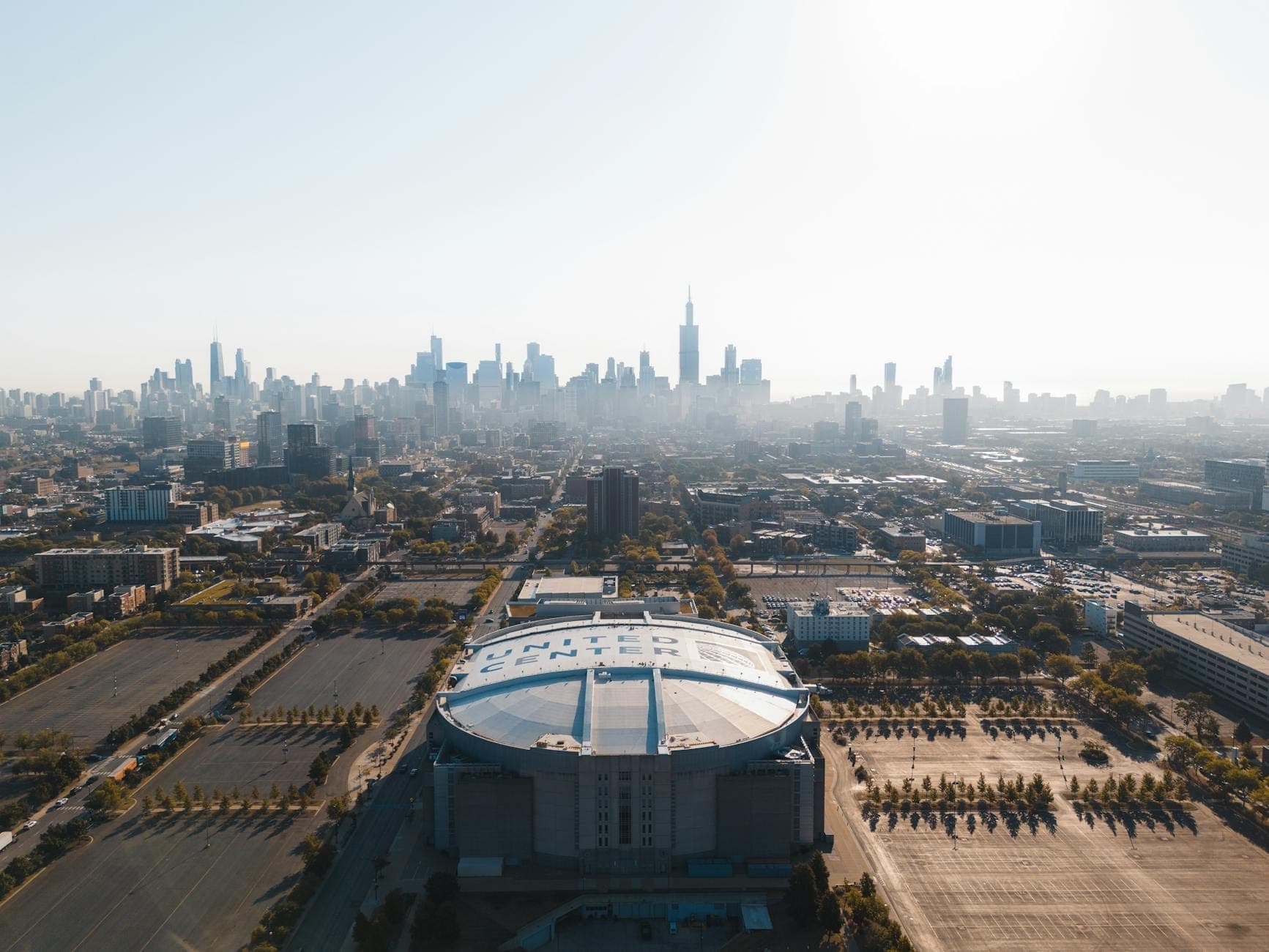 Aerial view of the United Center with the Chicago skyline in the background, showing the arena and its large parking lots on a clear day.