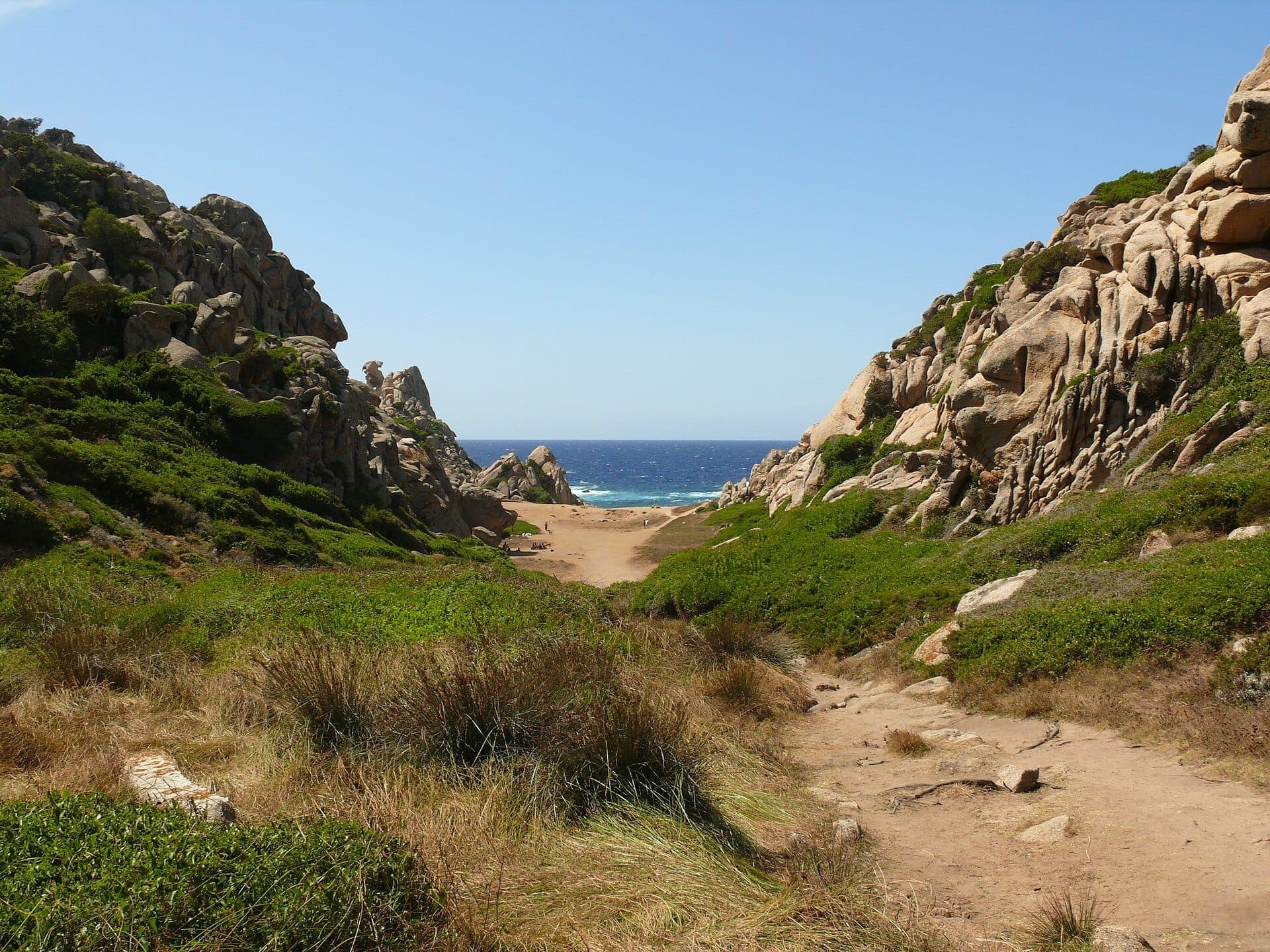 Vista del Valle della Luna, Capo Testa, con escarpados acantilados de granito, arbustos verdes, suelo arenoso del valle y el mar Mediterráneo azul al fondo.