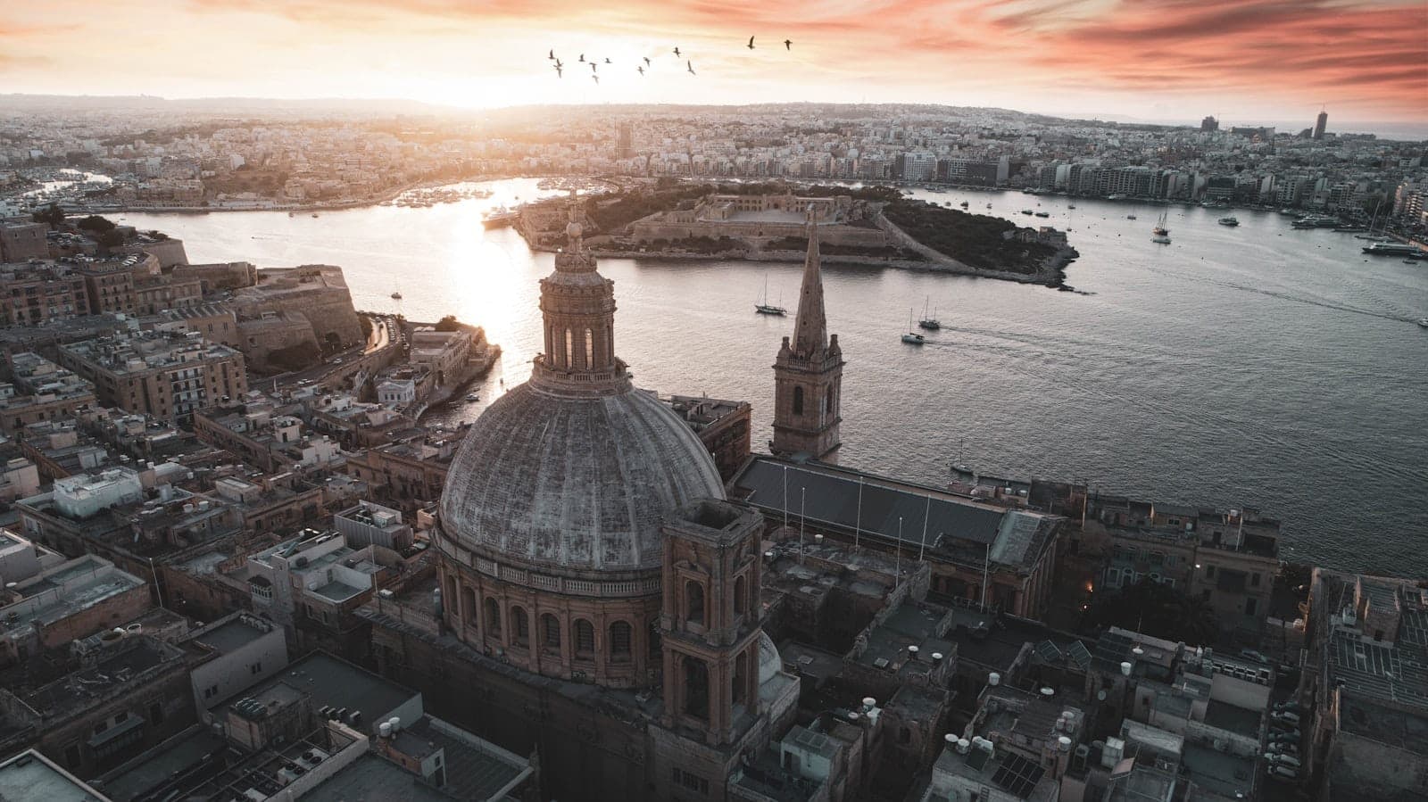 Atemberaubende Luftaufnahme von Valletta bei Sonnenuntergang: die ikonische Kuppel und der Kirchturm überragen den Grand Harbour, darunter Boote und ein dramatischer Himmel.