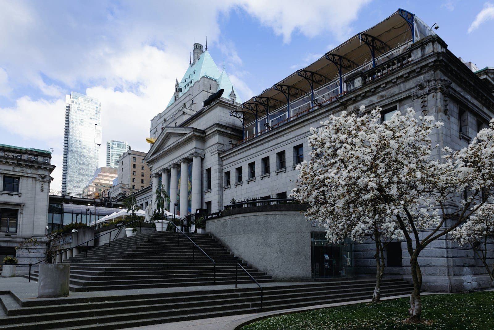 ด้านหน้าสไตล์นีโอคลาสสิกของ Vancouver Art Gallery พร้อมเสาขนาดใหญ่ ต้นไม้ดอกสีขาวบานสะพรั่ง และบันไดที่นำไปสู่อาคารสัญลักษณ์ในใจกลางเมืองภายใต้ท้องฟ้าที่มีเมฆบางส่วน