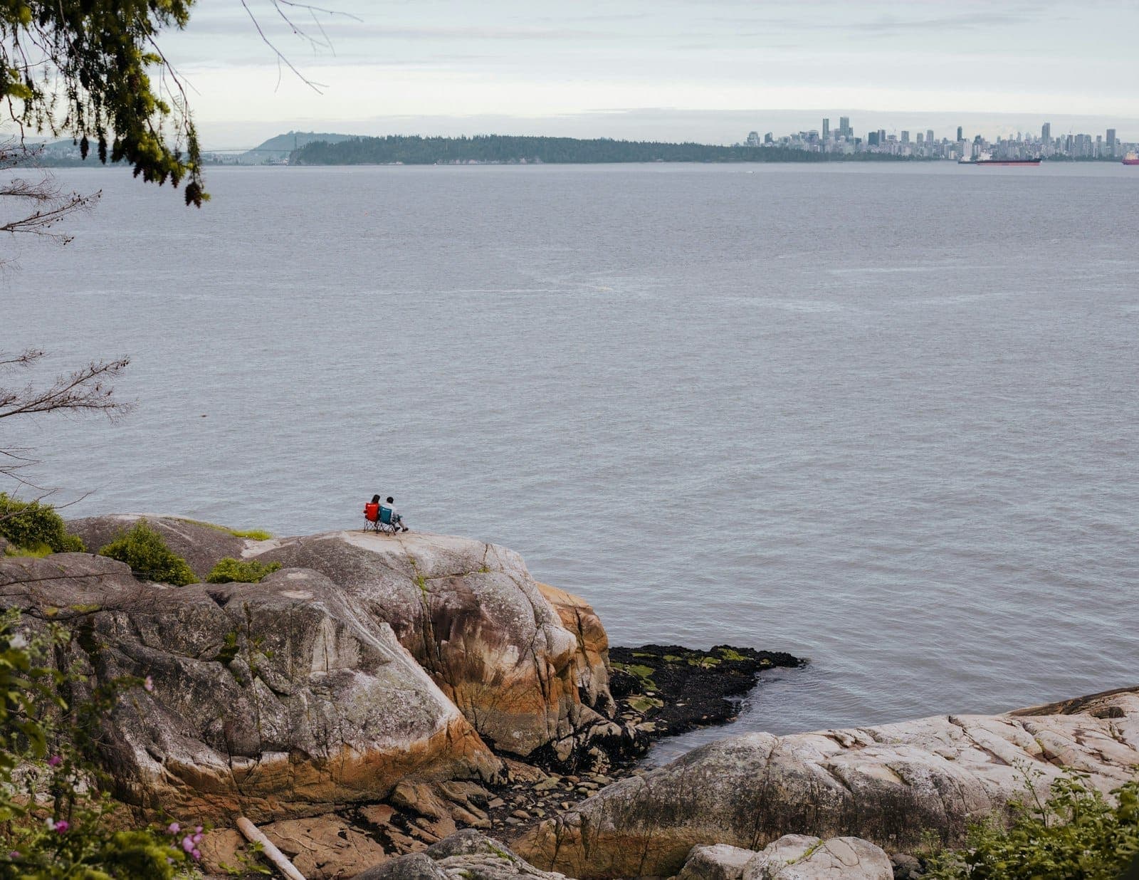 คู่รักนั่งบนโขดหินชมวิวทะเลพร้อมเส้นขอบฟ้าของ Vancouver ในระยะไกล ท่ามกลางทัศนียภาพชายฝั่งธรรมชาติ