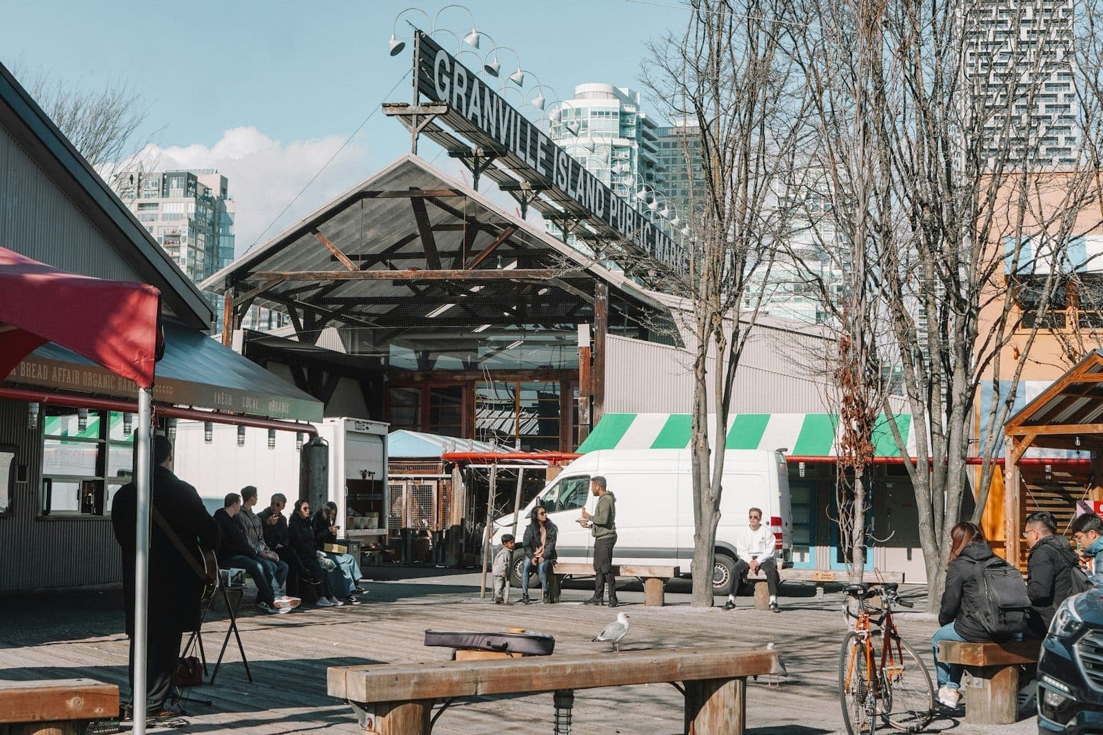 พื้นที่กลางแจ้งของ Granville Island Public Market พร้อมผู้คนนั่งบนม้านั่งและเส้นขอบฟ้าของ Vancouver เป็นฉากหลัง