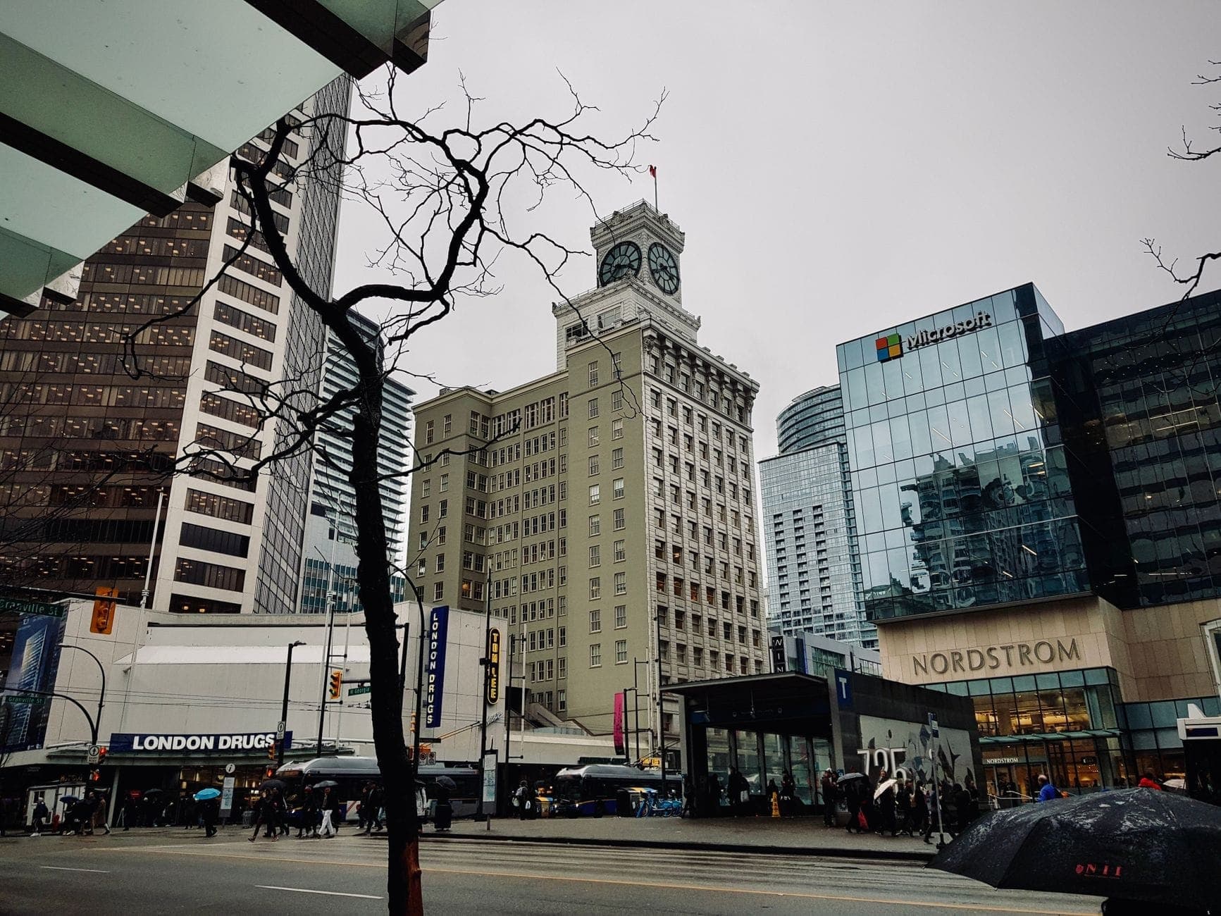 ใจกลางเมือง Vancouver ในวันที่มีฝนตกและท้องฟ้าครึ้ม มีผู้คนถือร่ม อาคารในเมือง และถนนที่เปียกชื้น