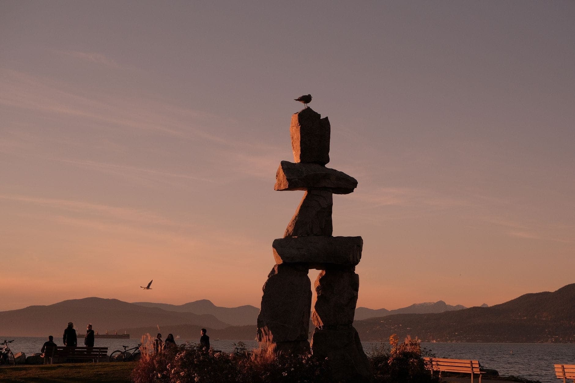 รูปปั้นหินอินุกชุก (Inukshuk) ใกล้ริมน้ำของแวนคูเวอร์ในช่วงพระอาทิตย์ตก พร้อมม้านั่ง น้ำ และภูเขาในพื้นหลัง