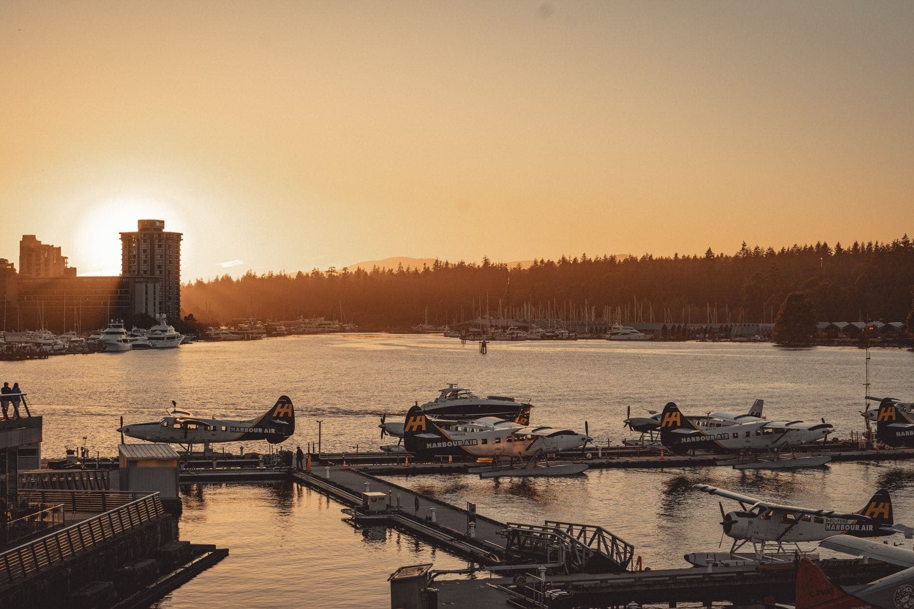 เครื่องบินทะเลของ Harbour Air หลายลำจอดเทียบท่าที่ท่าเรือ Vancouver ยามพระอาทิตย์ตก โดยมีเส้นขอบฟ้าของเมืองและ Stanley Park เป็นฉากหลัง