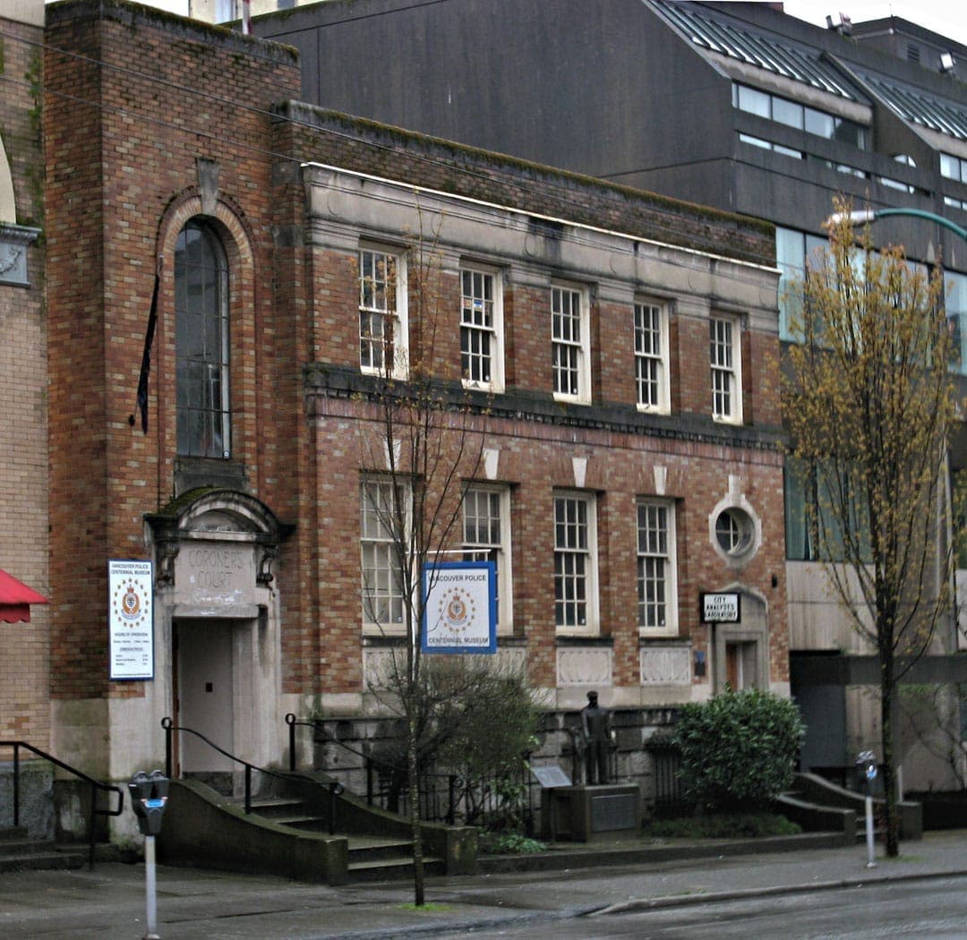 มุมมองภายนอกของอาคาร Vancouver Police Museum & Archives โดดเด่นด้วยผนังอิฐสีแดง หน้าต่างคลาสสิก ป้ายทางเข้า และแสงไฟถนนในวันที่ท้องฟ้าครึ้ม