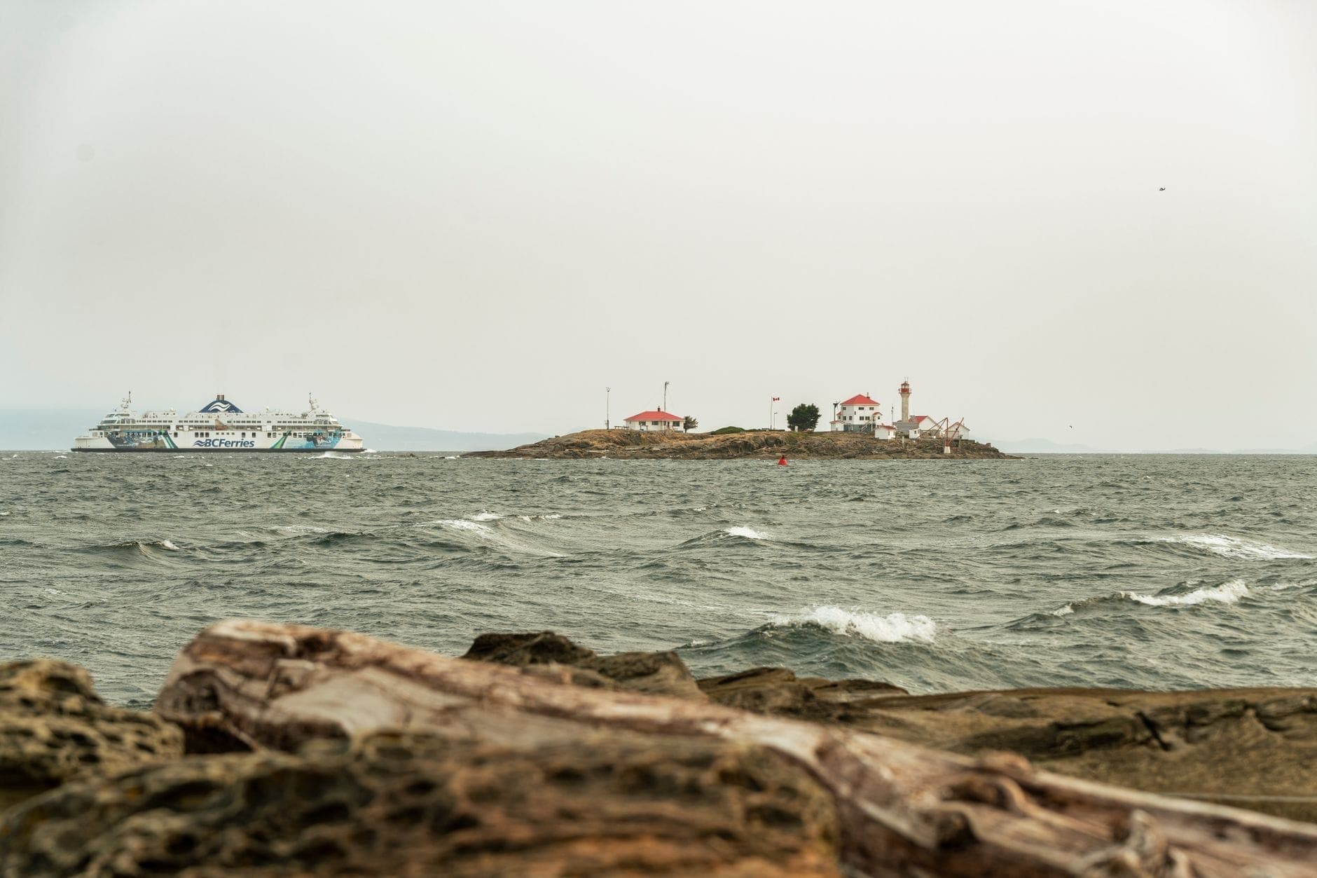 วิวเหนือผืนน้ำพร้อมเรือเฟอร์รี่ของ BC Ferries และประภาคารบนเกาะเล็กๆ ใต้ท้องฟ้าที่มีเมฆปกคลุม สื่อถึงการเดินทางระหว่าง Vancouver และ Vancouver Island