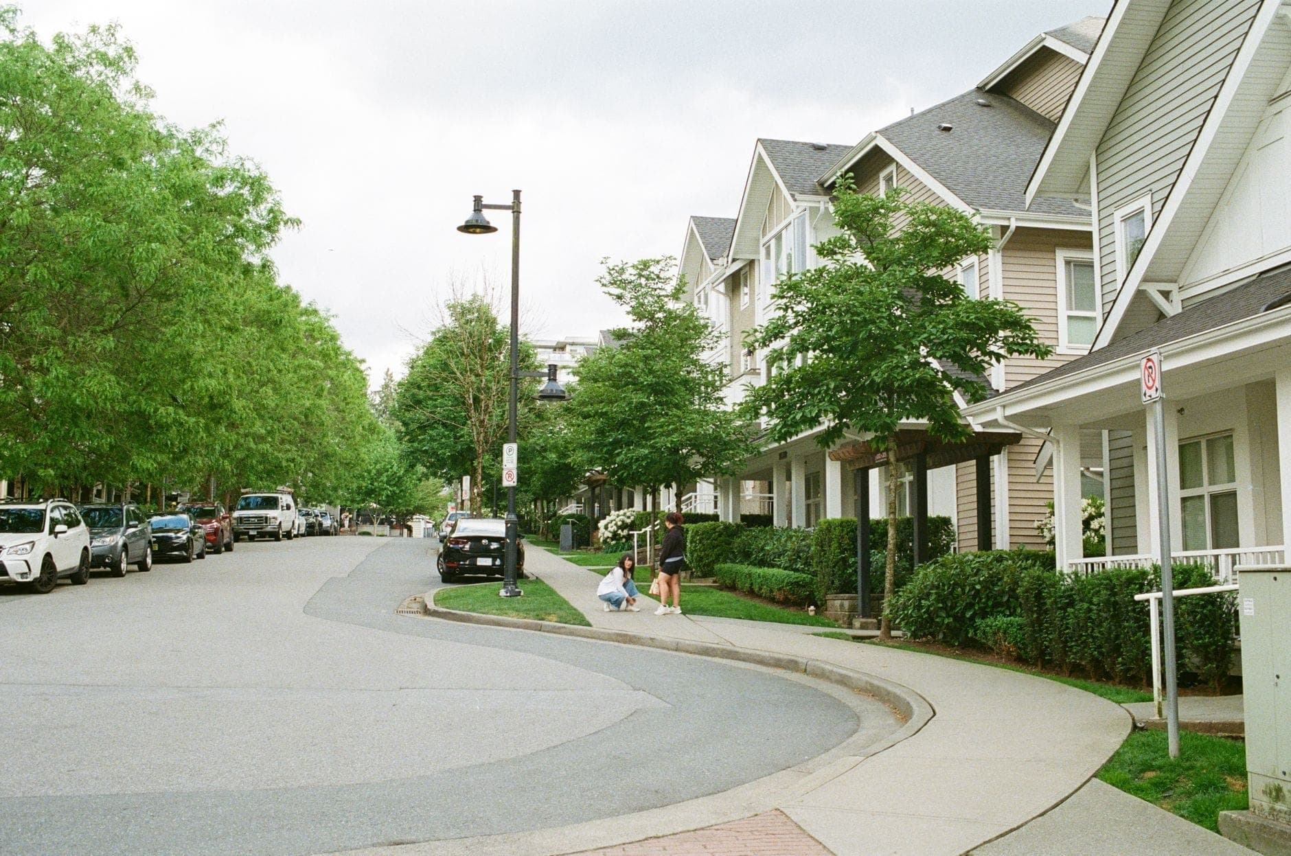 ย่านที่พักอาศัยที่เงียบสงบใน Vancouver พร้อมทาวน์เฮาส์ ต้นไม้เขียวชะอุ่ม รถจอดริมถนน และครอบครัวหนึ่งกำลังเดินบนทางเท้า