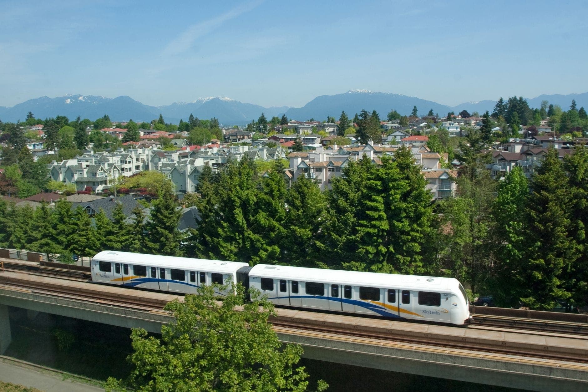 รถไฟ Vancouver SkyTrain วิ่งบนรางยกสูงเหนือย่านที่พักอาศัยสีเขียวขจี โดยมีภูเขาปรากฏให้เห็นในพื้นหลังใต้ท้องฟ้าที่แจ่มใส
