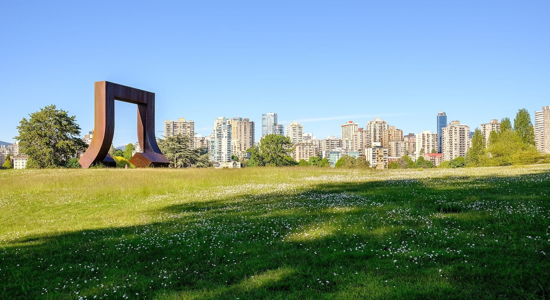 ทุ่งหญ้าสีเขียวกว้างใหญ่พร้อมดอกไม้ป่าใน Vanier Park ประติมากรรมศิลปะโลหะขนาดใหญ่ และตึกระฟ้าของ Vancouver ใต้ท้องฟ้าสีฟ้าแจ่มใส