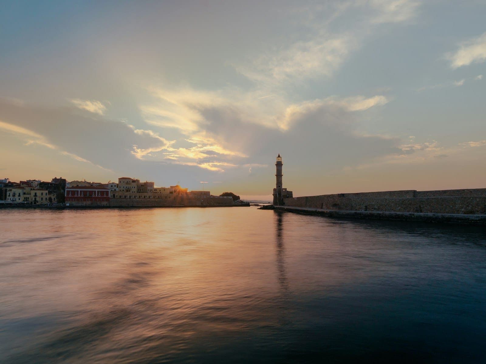 Sonnenuntergang über dem venezianischen Hafen von Chania mit dem ikonischen Leuchtturm und historischen Ufergebäuden, die sich in ruhigem, farbenprächtigen Wasser unter einem dramatischen Himmel spiegeln.