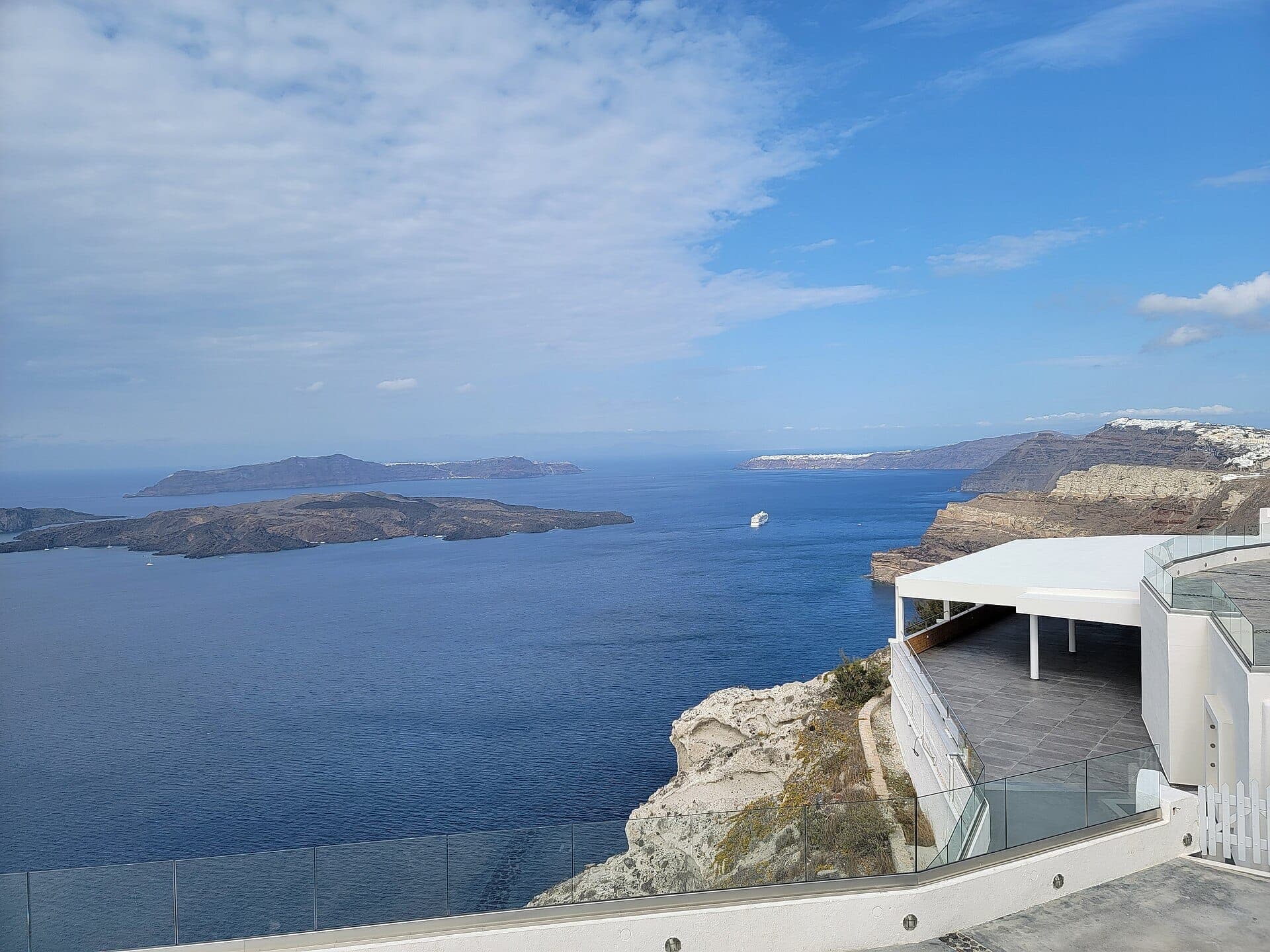 Die Terrasse der Venetsanos Winery mit Blick auf Santorinis blaue Caldera, steile Klippen, entfernte Inseln und ein weißes Kreuzfahrtschiff an einem hellen, sonnigen Tag.