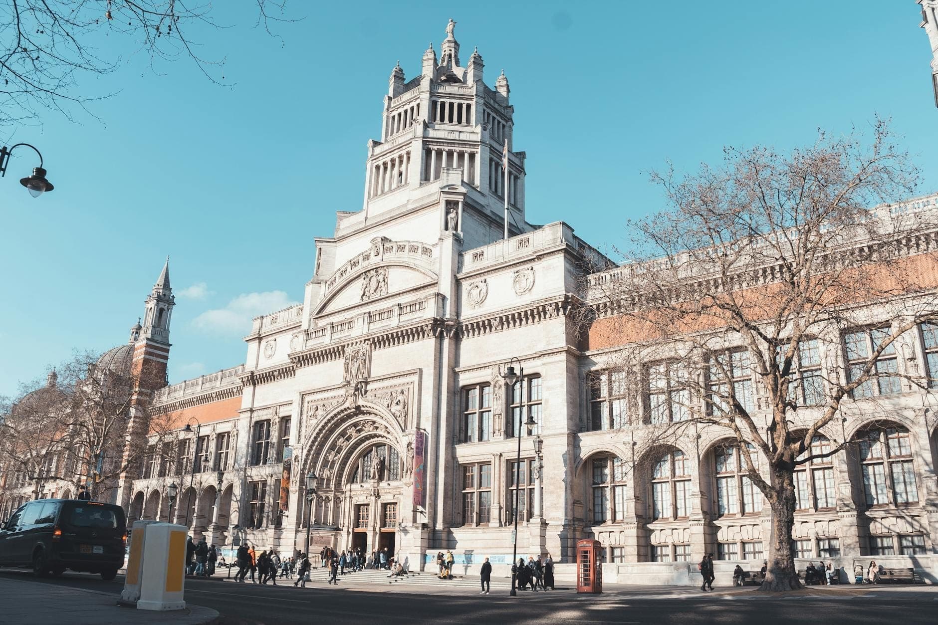 Weitwinkelansicht der imposanten Fassade des Victoria and Albert Museum mit Passanten davor unter einem klaren blauen Himmel in South Kensington, London.