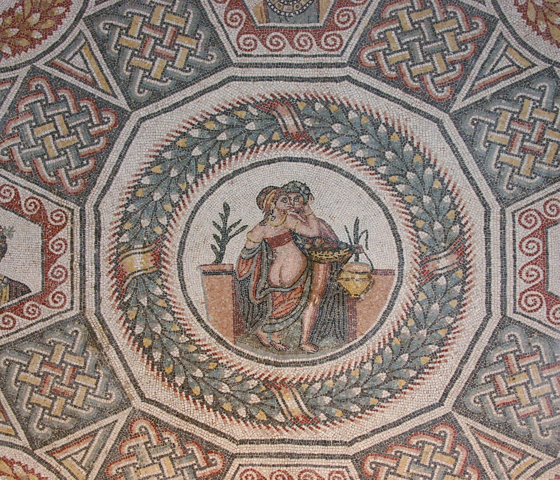 Lantai mosaik Romawi yang mendetail di Villa Romana del Casale, menampilkan pola geometris yang cerah dan adegan sentral dua figur yang dikelilingi karangan bunga laurel.