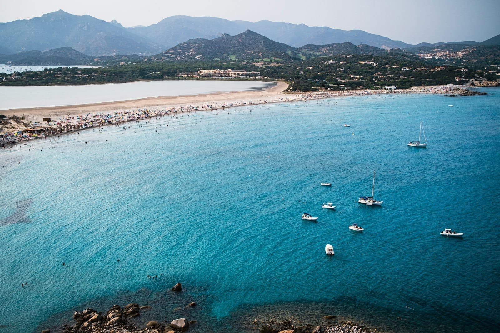 Vista aérea de una playa de arena blanca que se curva junto a aguas turquesas, con veleros anclados y montañas verdes al fondo, Villasimius, Cerdeña.