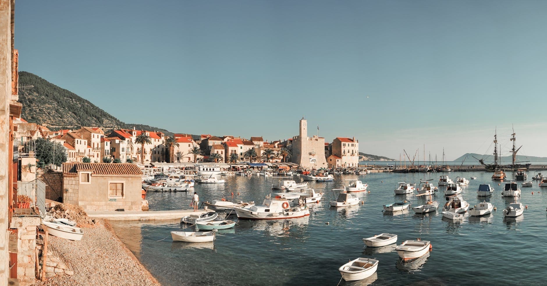 Vue panoramique du port de Vis avec des bateaux flottant dans des eaux cristallines, des bâtiments en pierre médiévaux et une tour sous un ciel bleu.
