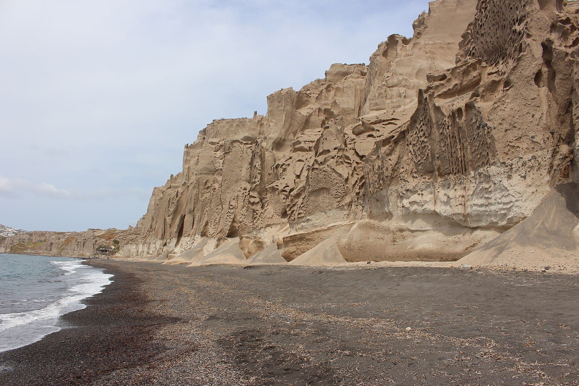 Hohe, erodierte Bimssteinklippen erheben sich über dem dunklen Sand und der Küstenlinie des Vlychada Beach an Santorinis dramatischer Südküste unter einem bewölkten Himmel.