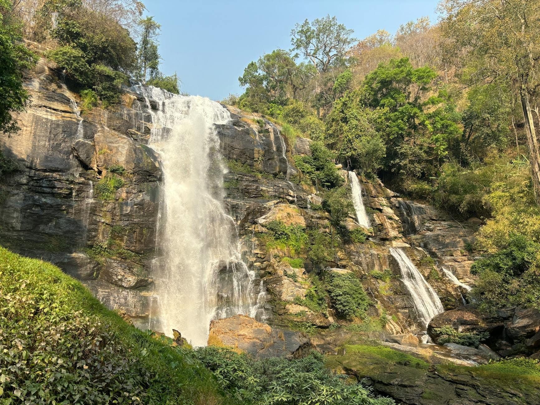 Der Wachirathan-Wasserfall stürzt über eine felsige Klippe inmitten üppigen grünen Waldes unter blauem Himmel im Doi-Inthanon-Nationalpark.